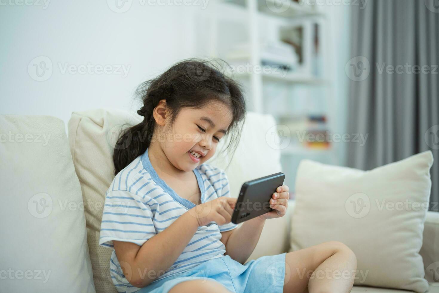 Asian child girl looking using and touch mobile phone screen on couch sofa. Baby smiling funny time to use mobile phone. Too much screen time. Cute girl watching videos while tv, Internet addiction. photo