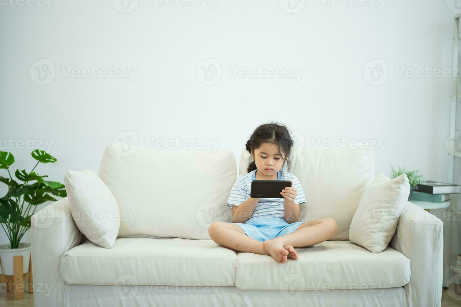 Asian child girl looking using and touch mobile phone screen on couch sofa. Baby smiling funny time to use mobile phone. Too much screen time. Cute girl watching videos while tv, Internet addiction. photo