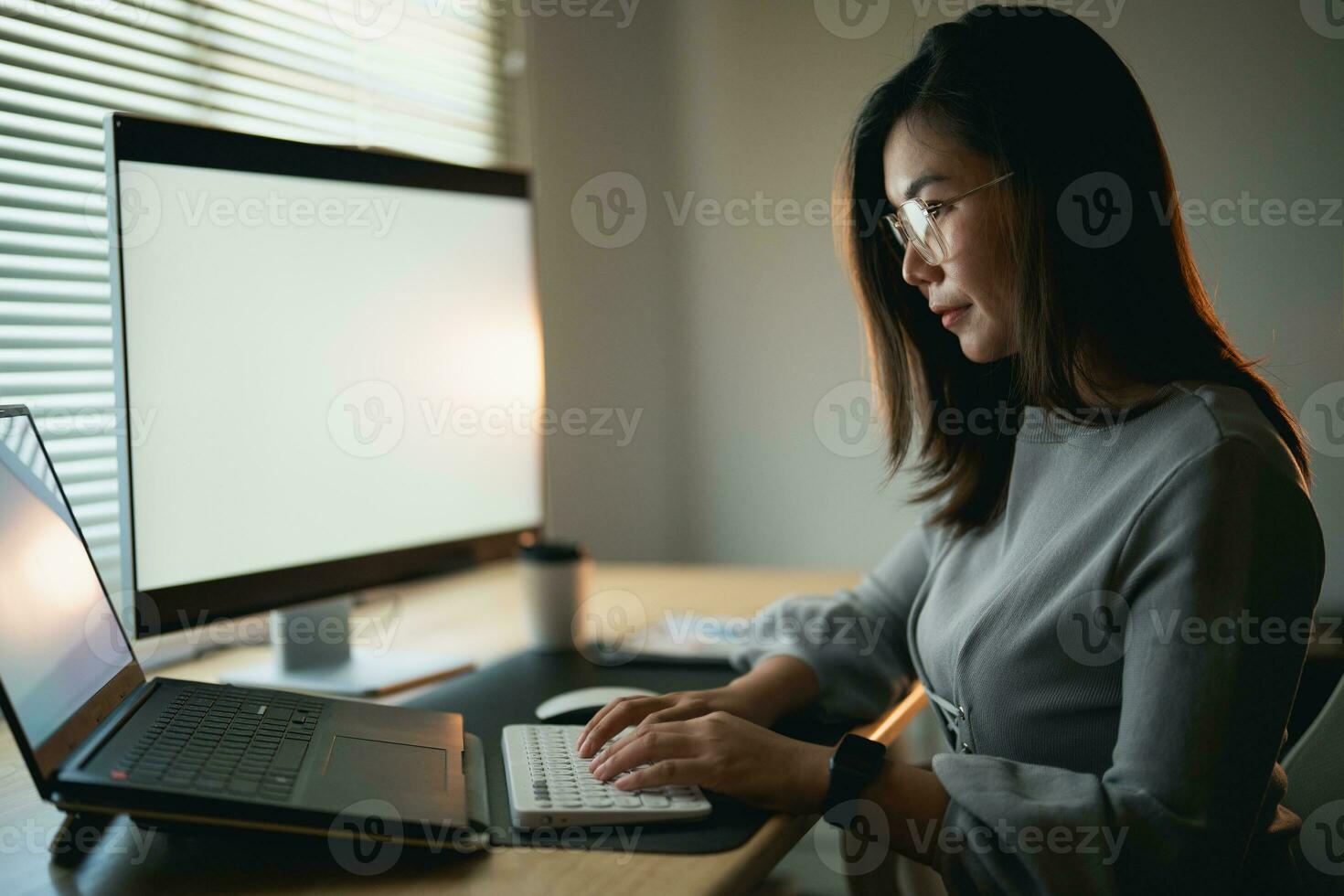 Asian businesswoman wearing suit and glasses typing on keyboard and working laptop on wooden table at home. Entrepreneur woman working for her business at living room home. Business work home concept. photo