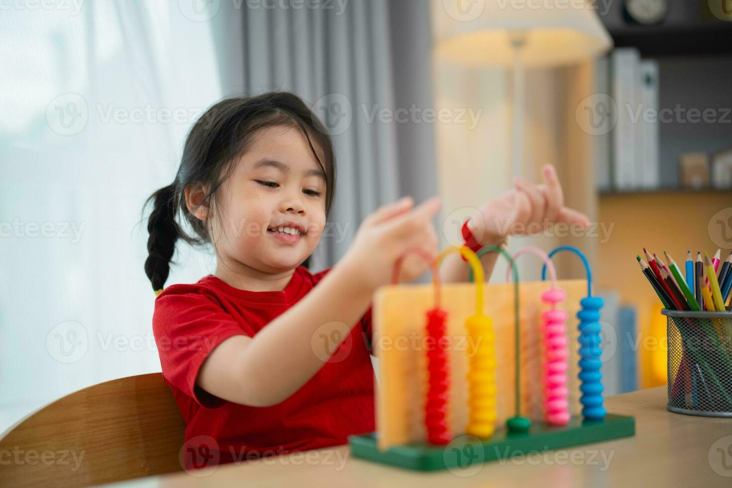 Young cute Asian baby girl wearing red t-shirt is learning the abacus with colored beads to learn how to count on the table in the living room at home. Child baby girl development studying concept. photo