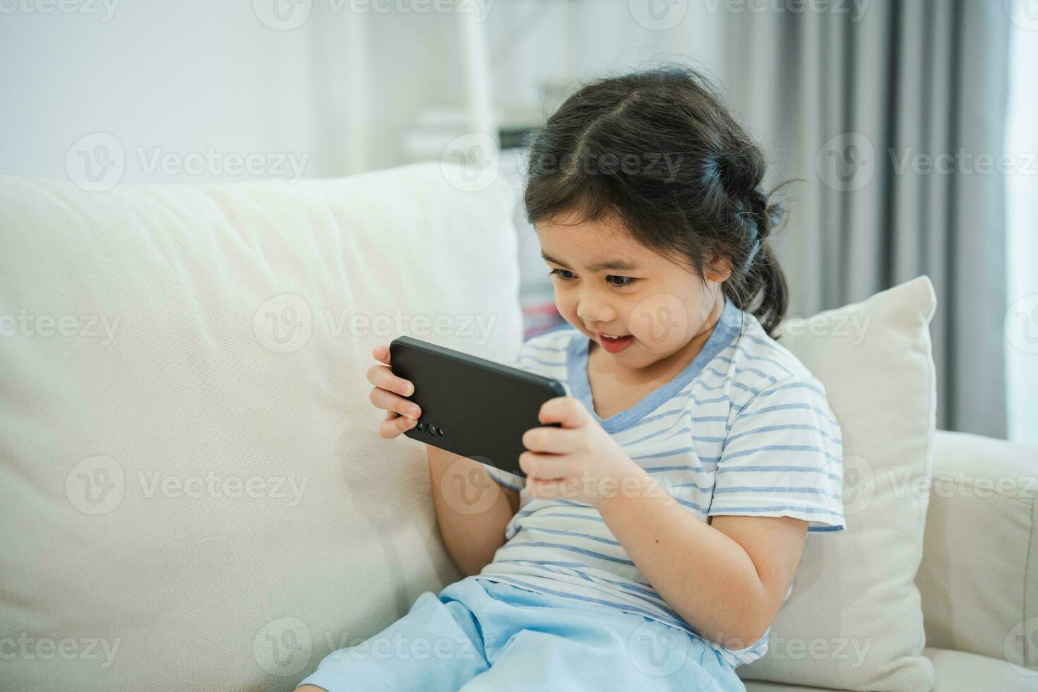 Asian child girl looking using and touch mobile phone screen on couch sofa. Baby smiling funny time to use mobile phone. Too much screen time. Cute girl watching videos while tv, Internet addiction. photo