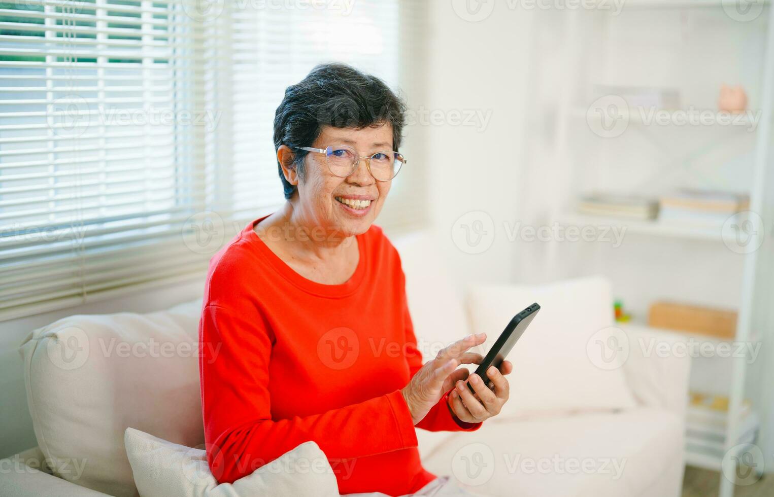 Happy senior old asian woman enjoying using mobile apps texting typing messages sit on sofa, smiling old lady holding smartphone looking smartphone browsing social media or learning technology at home photo