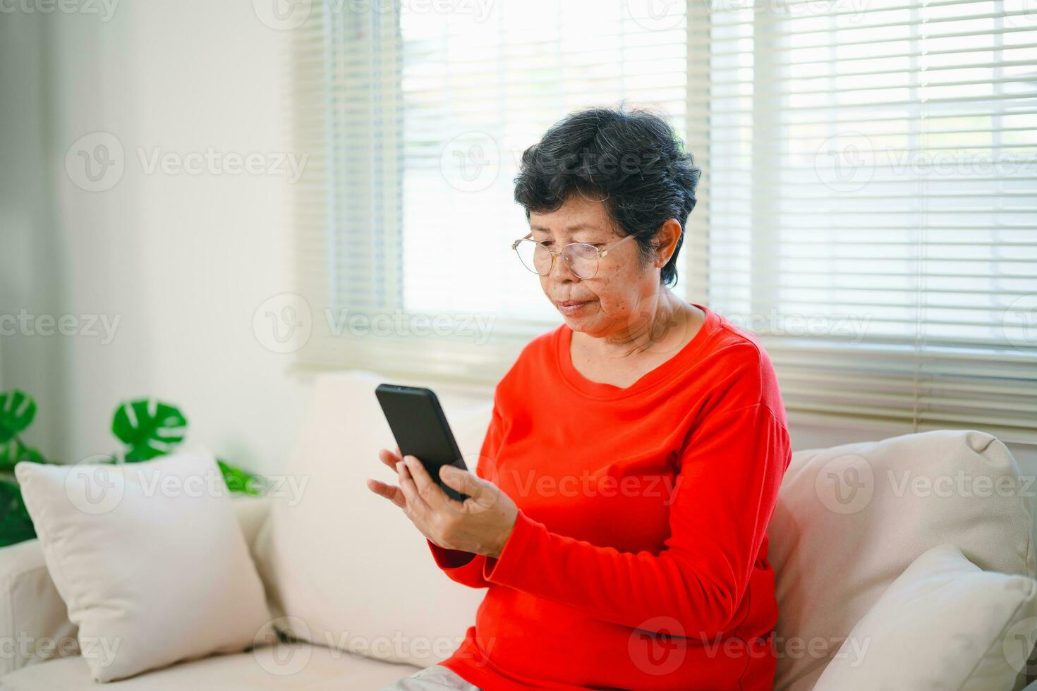 Happy senior old asian woman enjoying using mobile apps texting typing messages sit on sofa, smiling old lady holding smartphone looking smartphone browsing social media or learning technology at home photo
