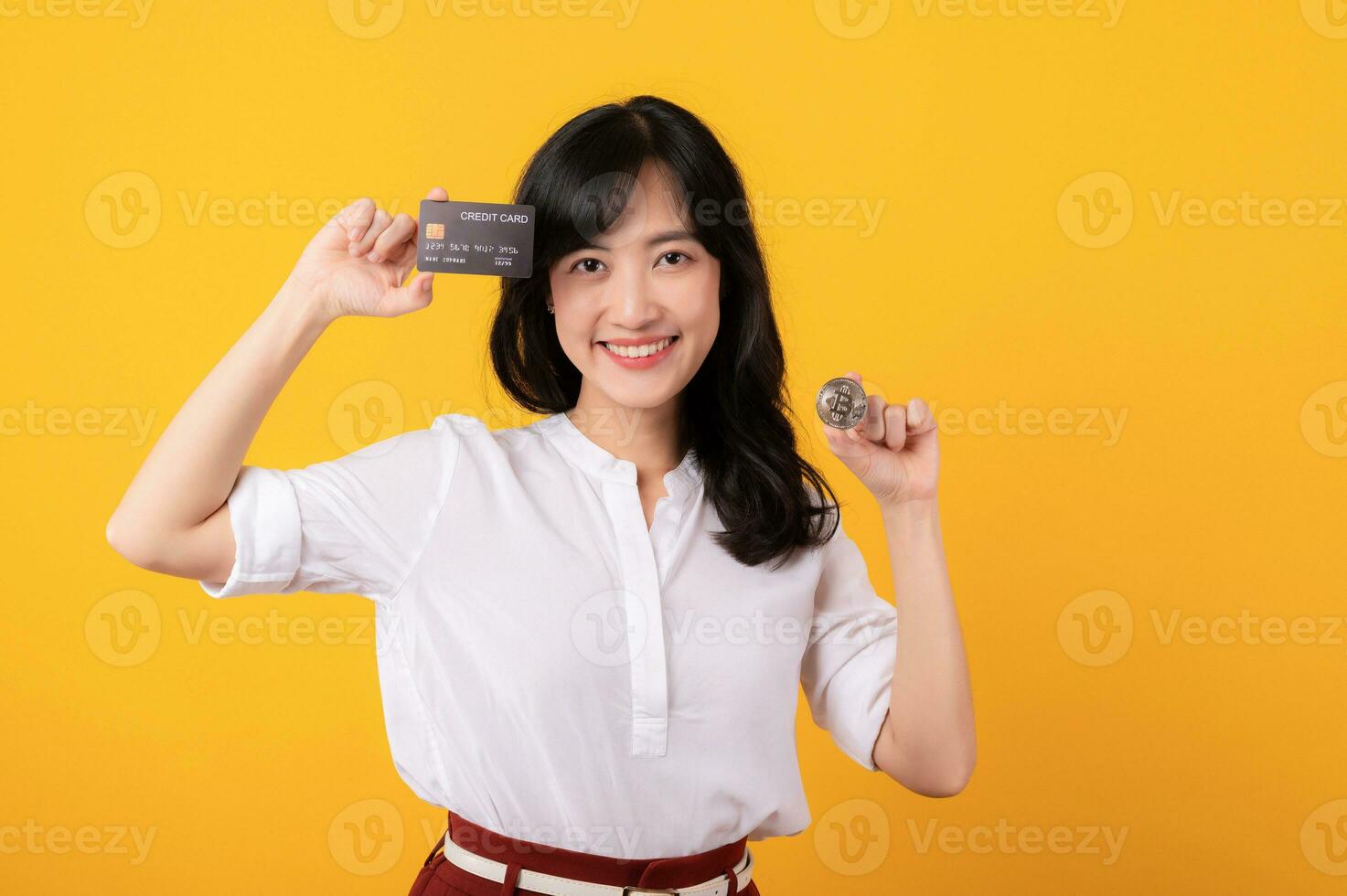 Portrait beautiful young asian woman enterpriser happy smile wearing white shirt and red plants holding credit card and crypto digital currency isolation on yellow background. Wealth concept. photo
