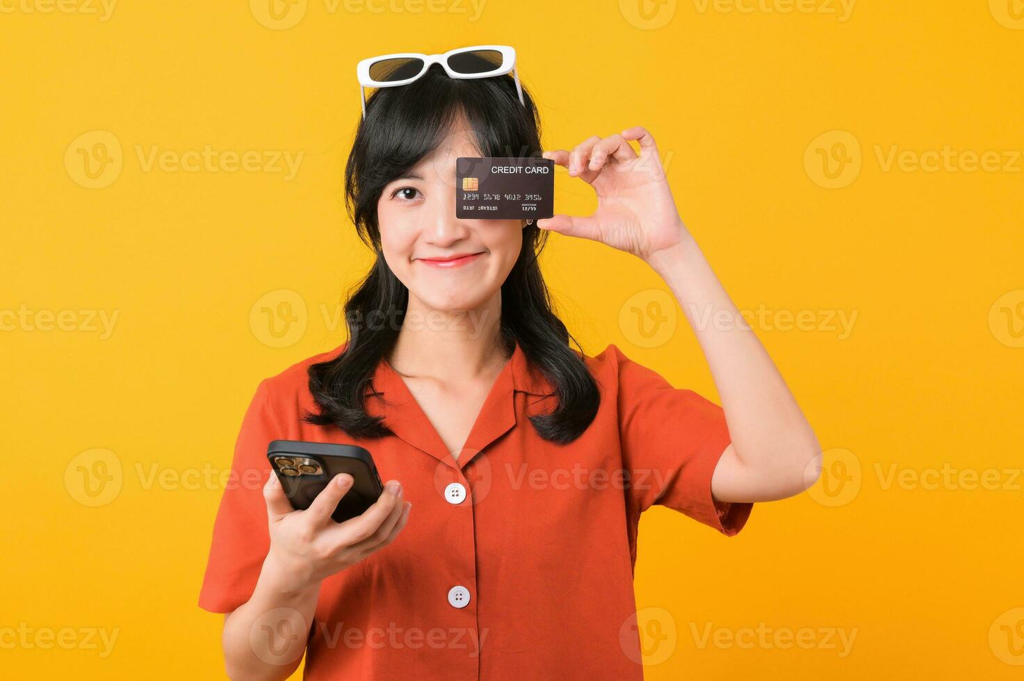 Portrait young beautiful asian woman happy smile dressed in orange clothes using smartphone and showing credit card isolated on yellow background. Pay and purchase shopping payment concept. photo