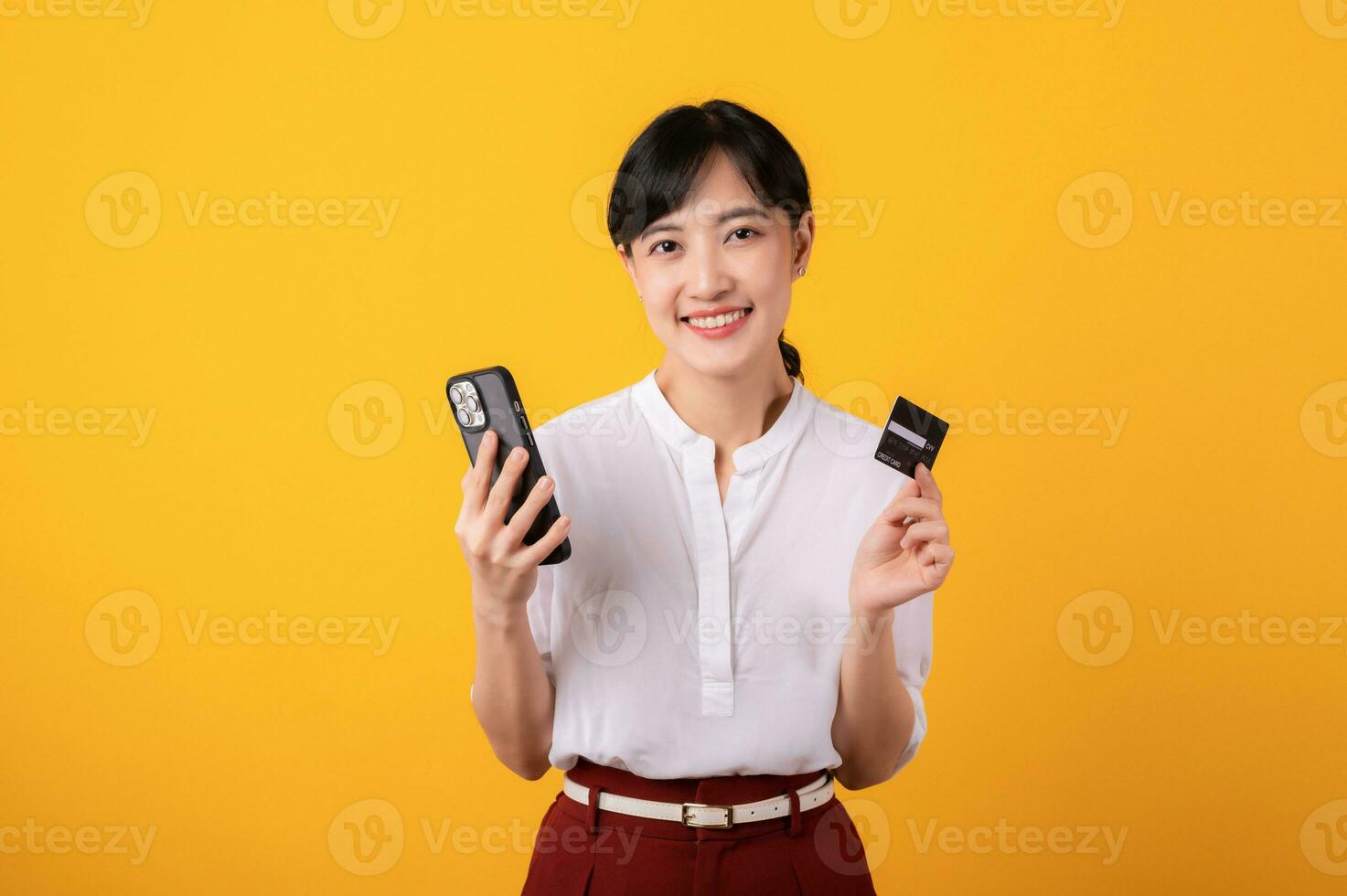 Portrait beautiful young asian woman enterpriser happy smile wearing white shirt and red plants holding credit card and smartphone isolation on yellow background. Online shopping concept. photo