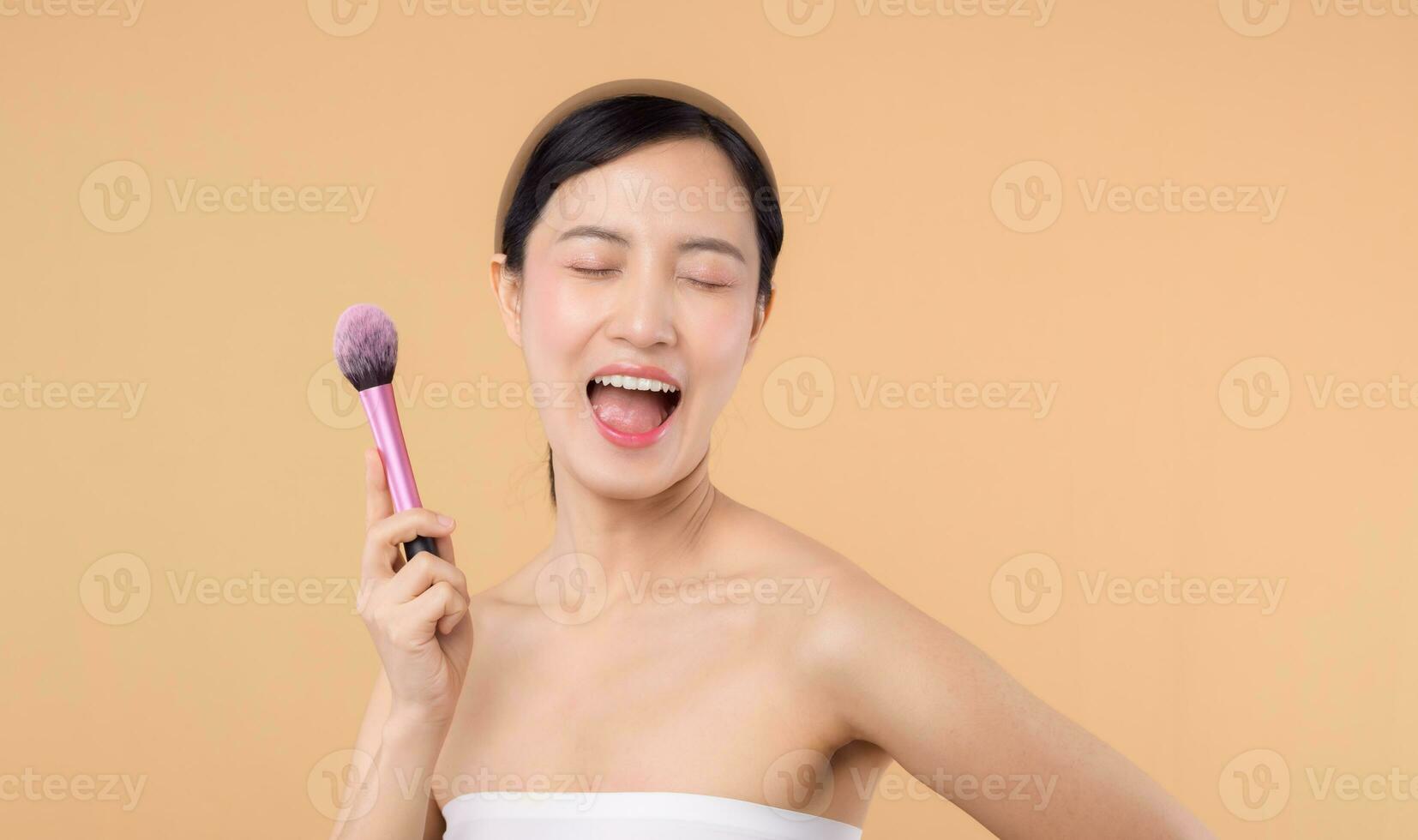 Portrait happy young asian woman with natural make up face holding cosmetic skin powder blusher isolated on beige background. Female apply skincare brush treatment. beauty product cosmetology concept. photo