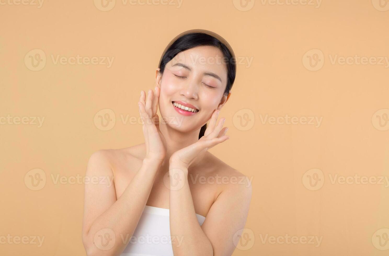 Beautiful girl asian model touching fresh glowing hydrated facial skin on beige background closeup. Beauty face young woman with natural makeup and healthy skin portrait. Skin care concept photo