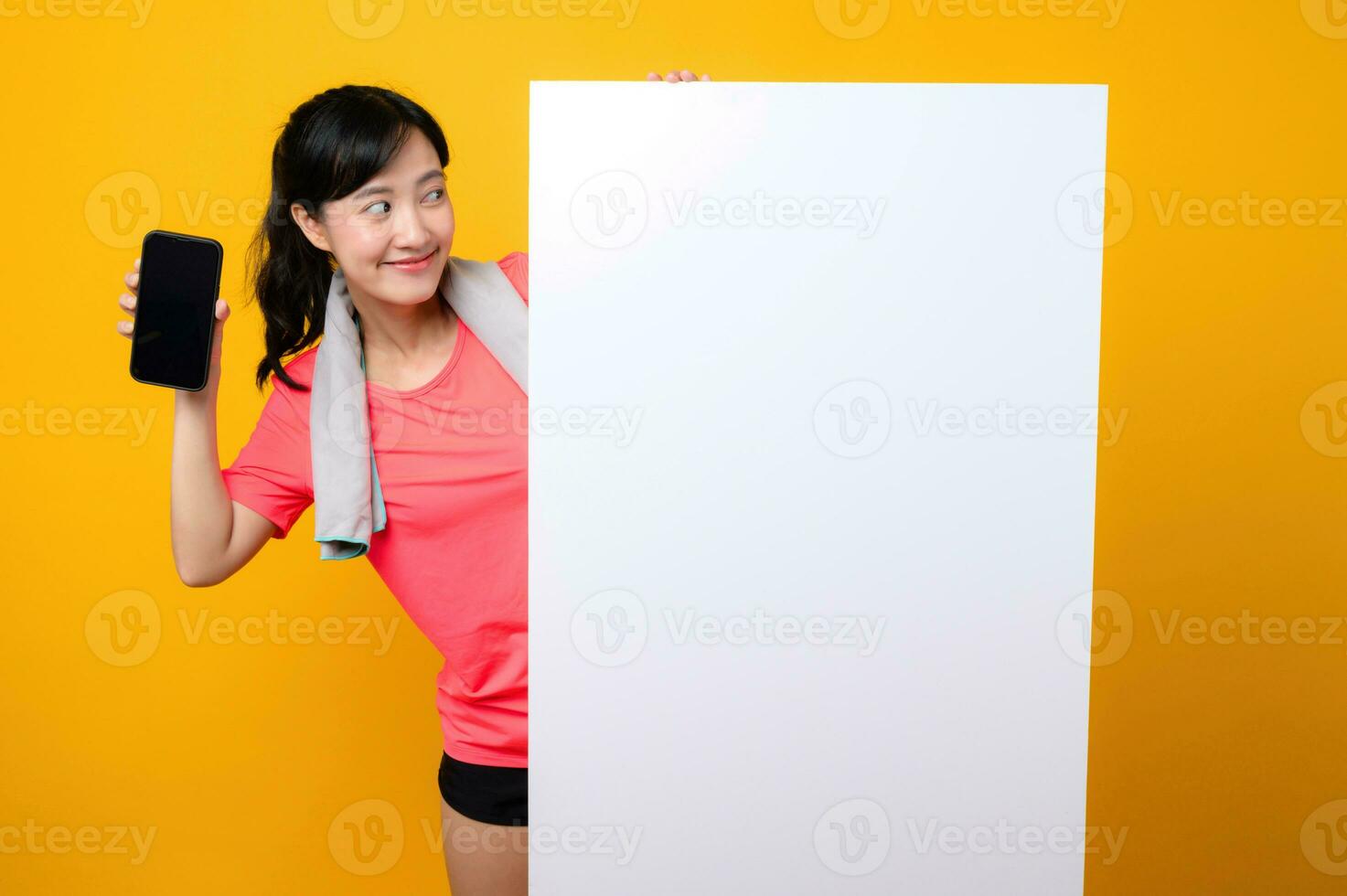 young asian sports fitness woman happy smile wearing pink sportswear standing behind the white blank banner or empty space advertisement board against yellow background. wellbeing application concept. photo