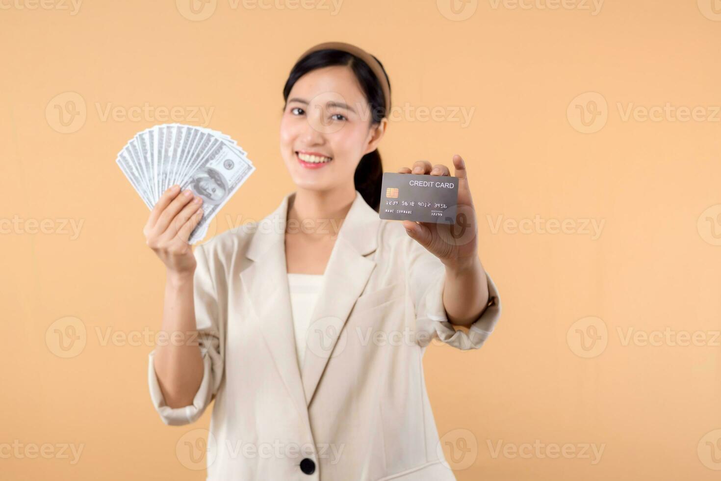 portrait of happy successful confident young asian business woman wearing white jacket holding cash money dollars and credit card standing over beige background. millionaire business, shopping concept photo