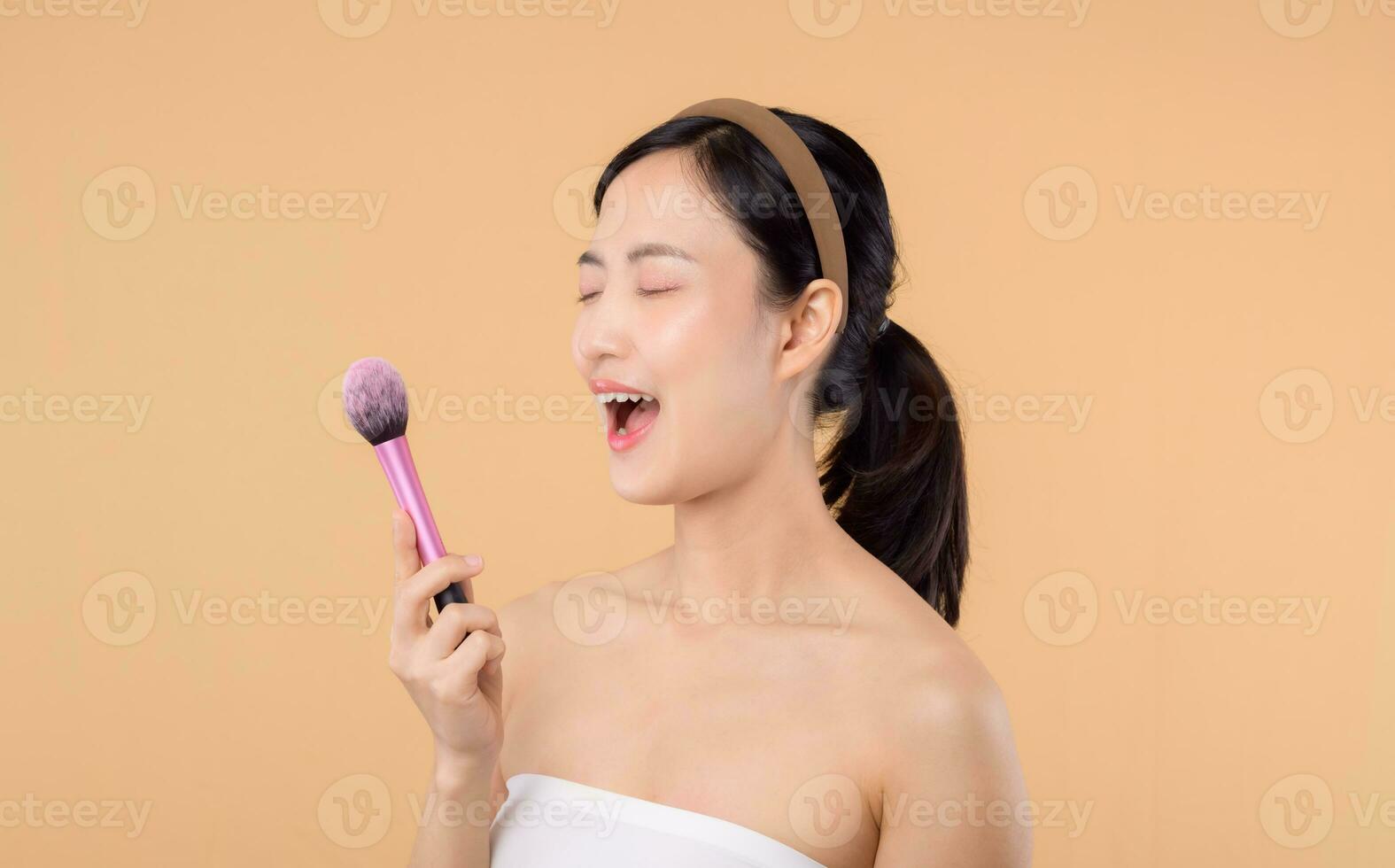 Portrait happy young asian woman with natural make up face holding cosmetic skin powder blusher isolated on beige background. Female apply skincare brush treatment. beauty product cosmetology concept. photo