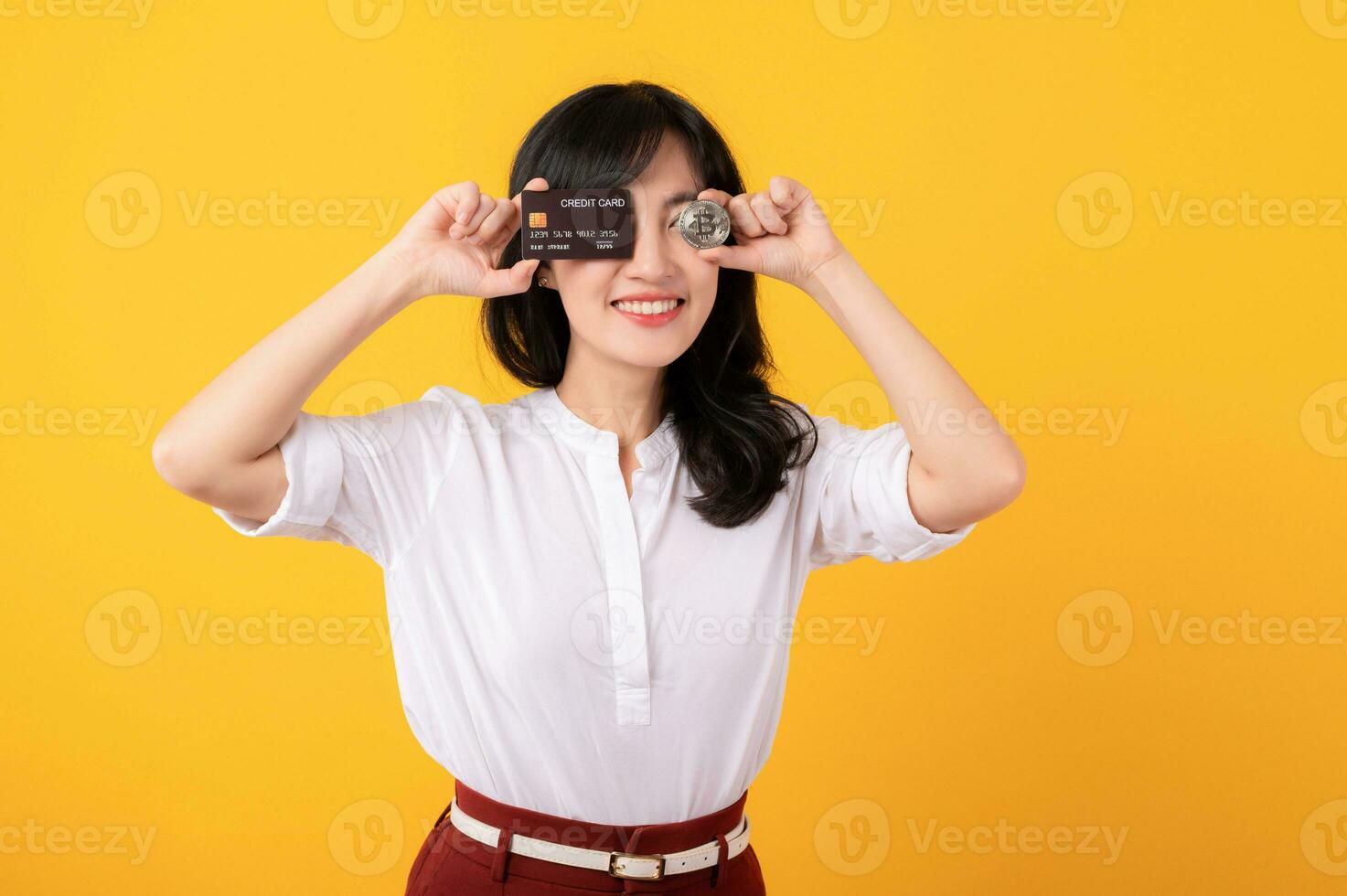 Portrait beautiful young asian woman enterpriser happy smile wearing white shirt and red plants holding credit card and crypto digital currency isolation on yellow background. Wealth concept. photo