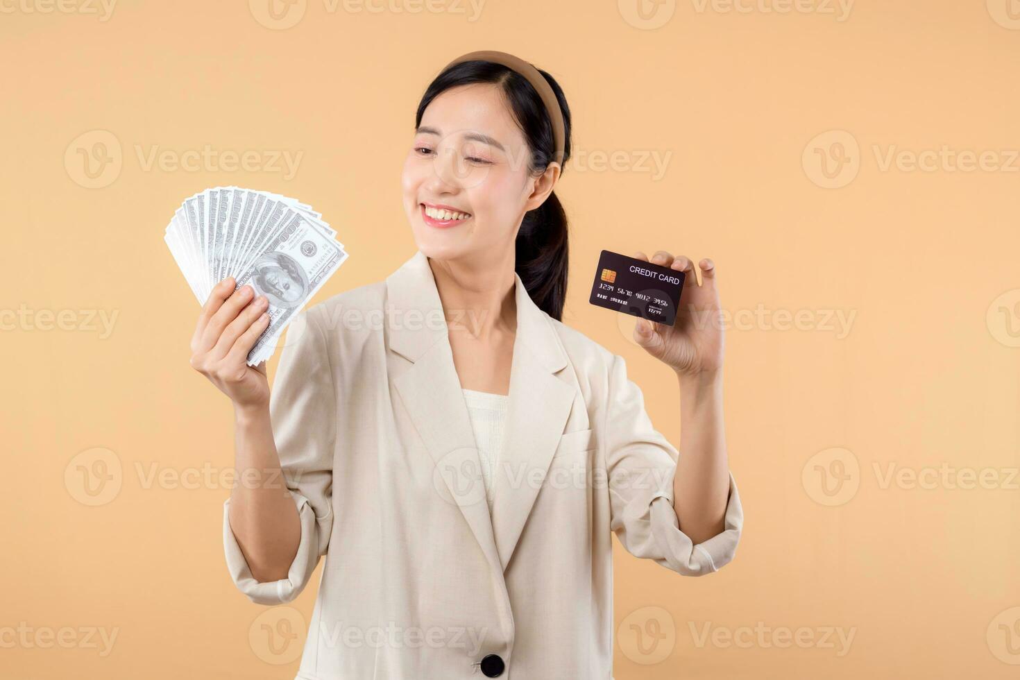 portrait of happy successful confident young asian business woman wearing white jacket holding cash money dollars and credit card standing over beige background. millionaire business, shopping concept photo