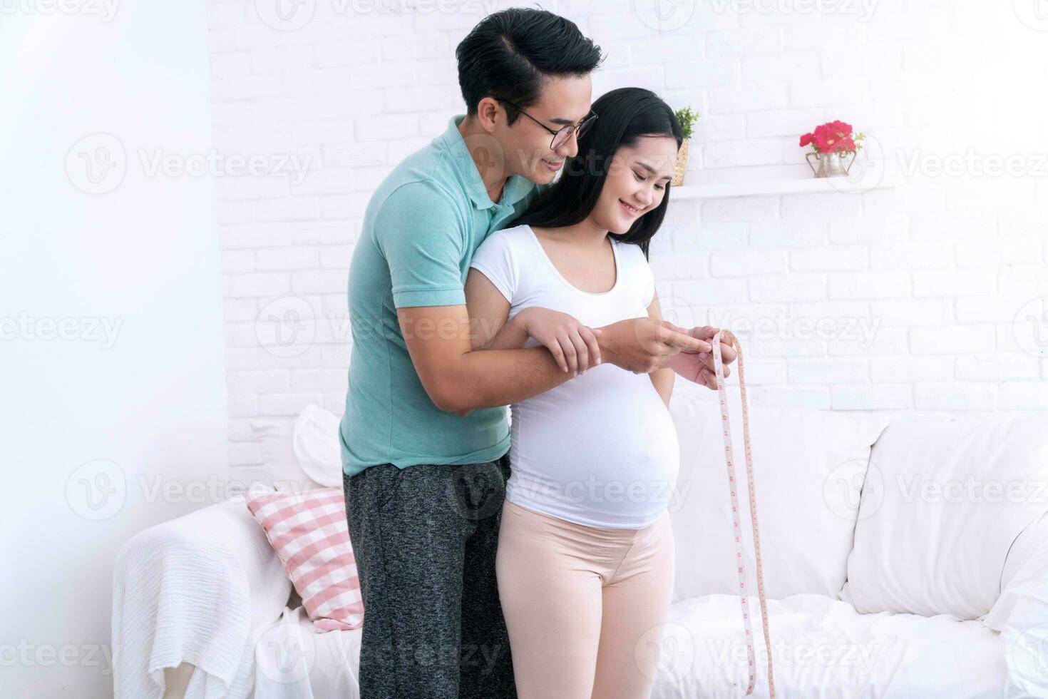 Happy young man measuring the waist of the pregnant his wife standing in living room. Husband measuring waist circumference his wife belly at home. photo