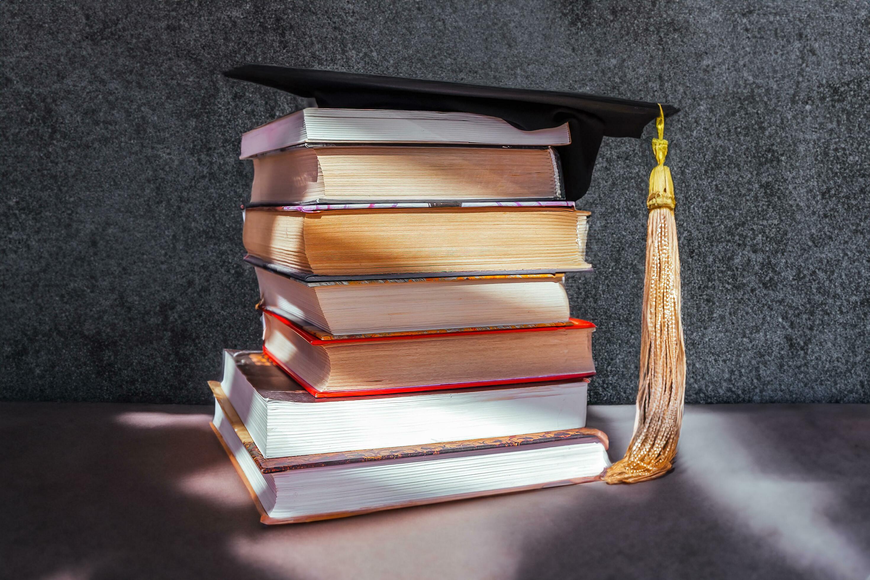 An Academic Cap Placed Atop A Stack Of Books Against A Wooden an-academic-cap-placed-atop-a-stack-of-books-against-a-wooden
