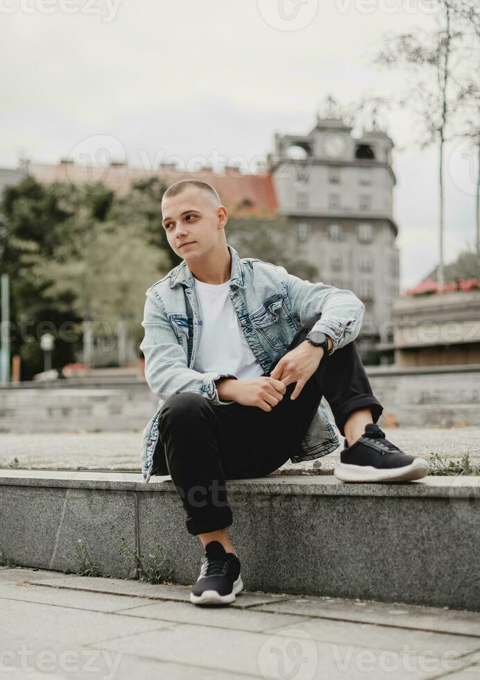 ciudad soledad. pensativo joven hombre en un mezclilla chaqueta. elegante melancólico joven chico reflexiona vida en su mezclilla chaqueta. paisaje urbano contemplación. joven hombre en mezclilla chaqueta perdido en pensamiento foto