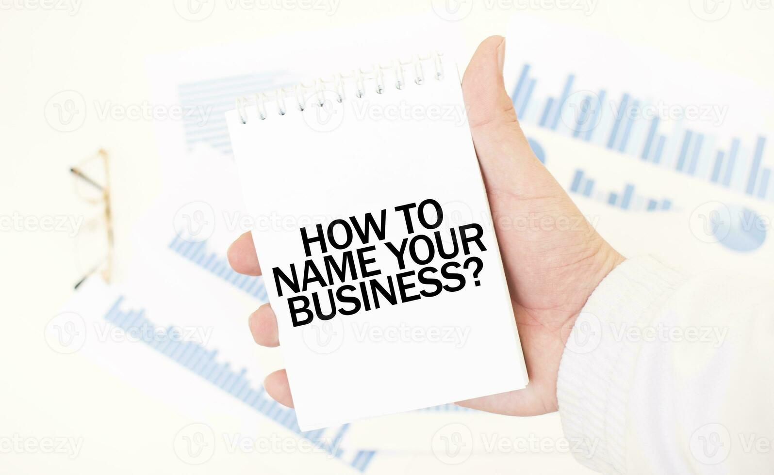 Businessman holding a white notepad with text HOW TO NAME YOUR BUISINESS, business concept photo