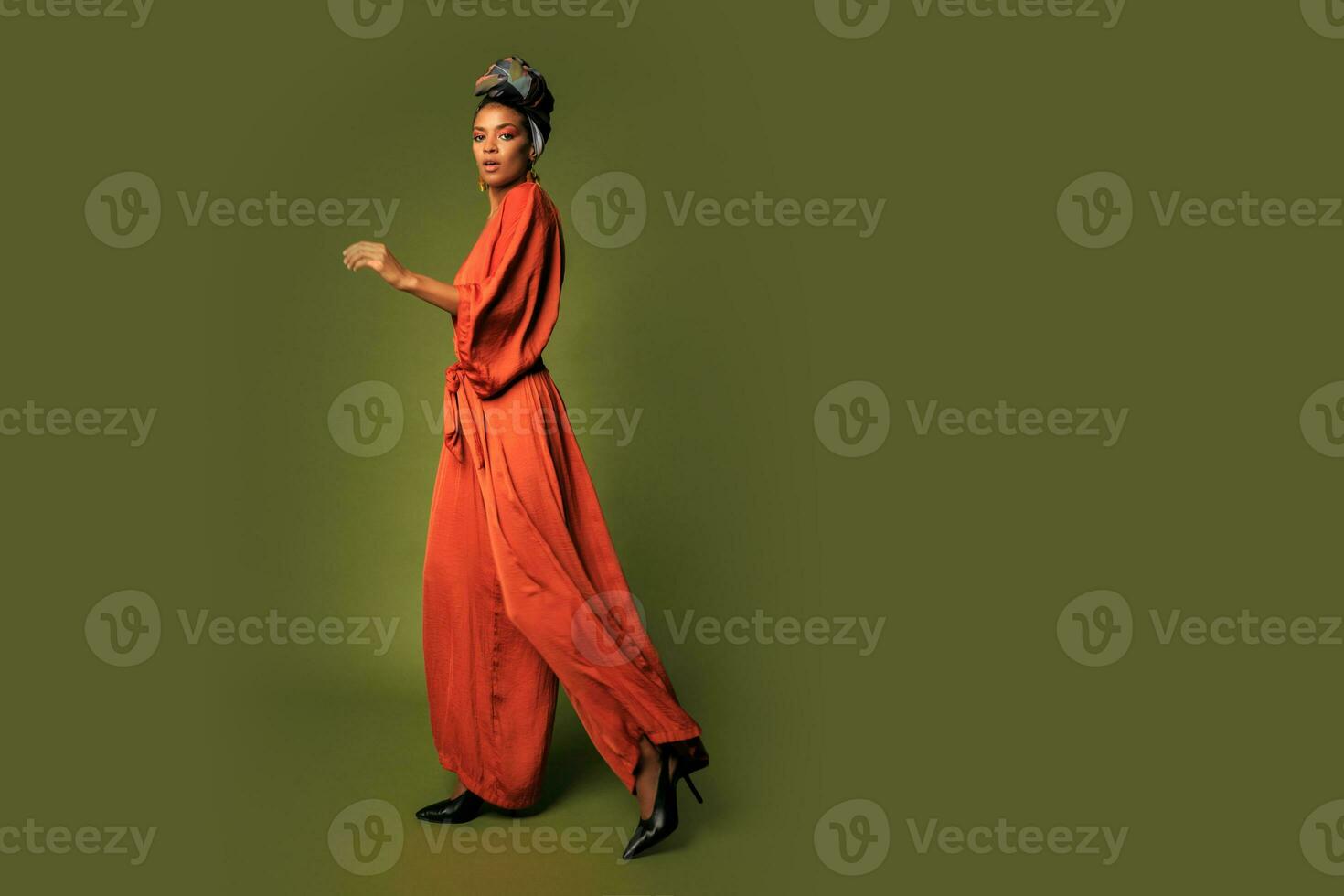 Fashion studio photo of stylish african woman with turban and orange dress posing  over green background.