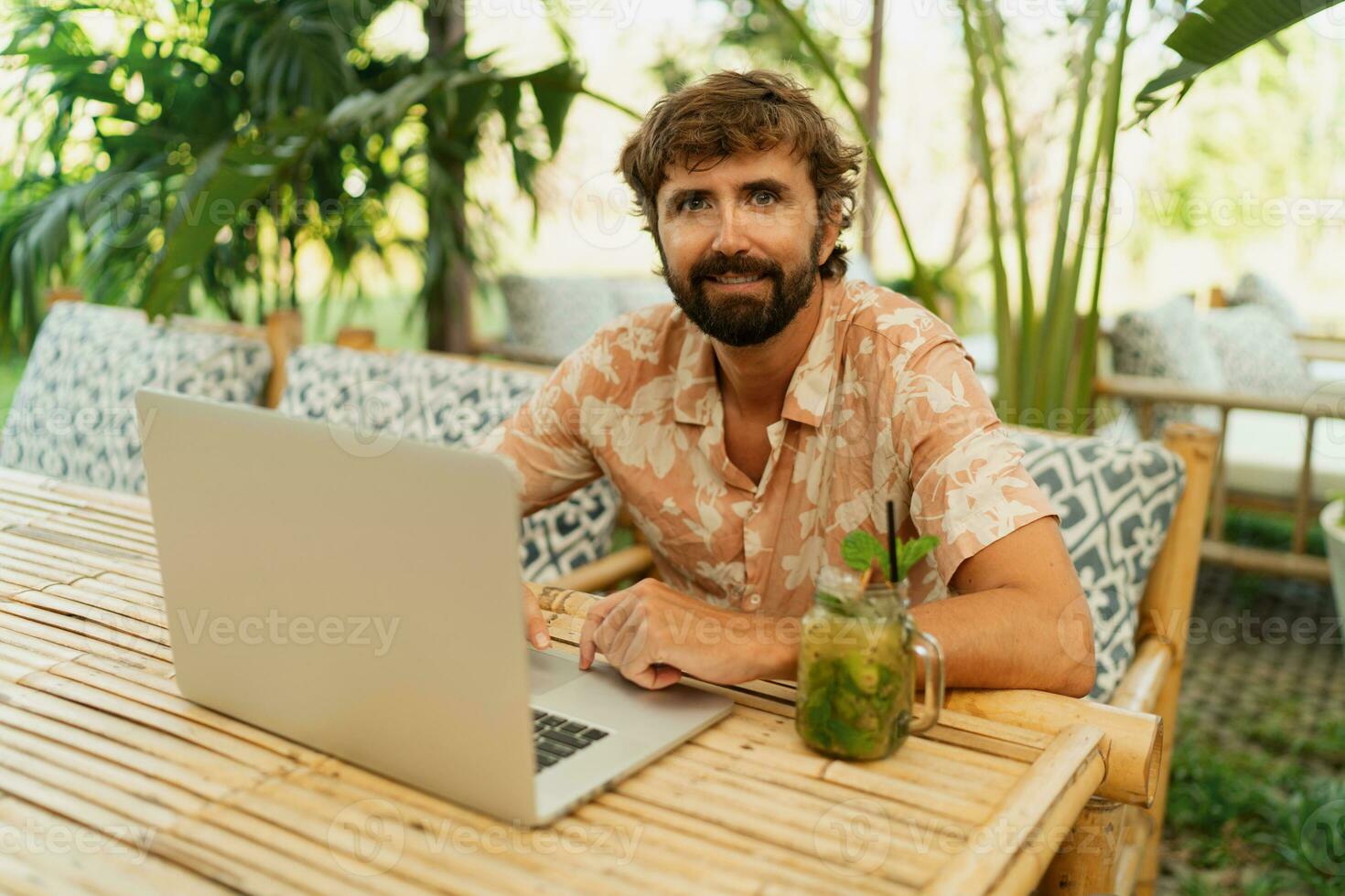 Handsome man with beard using lap top and mobyle phone , sitting in outdoor  cafe with tropical interior. photo