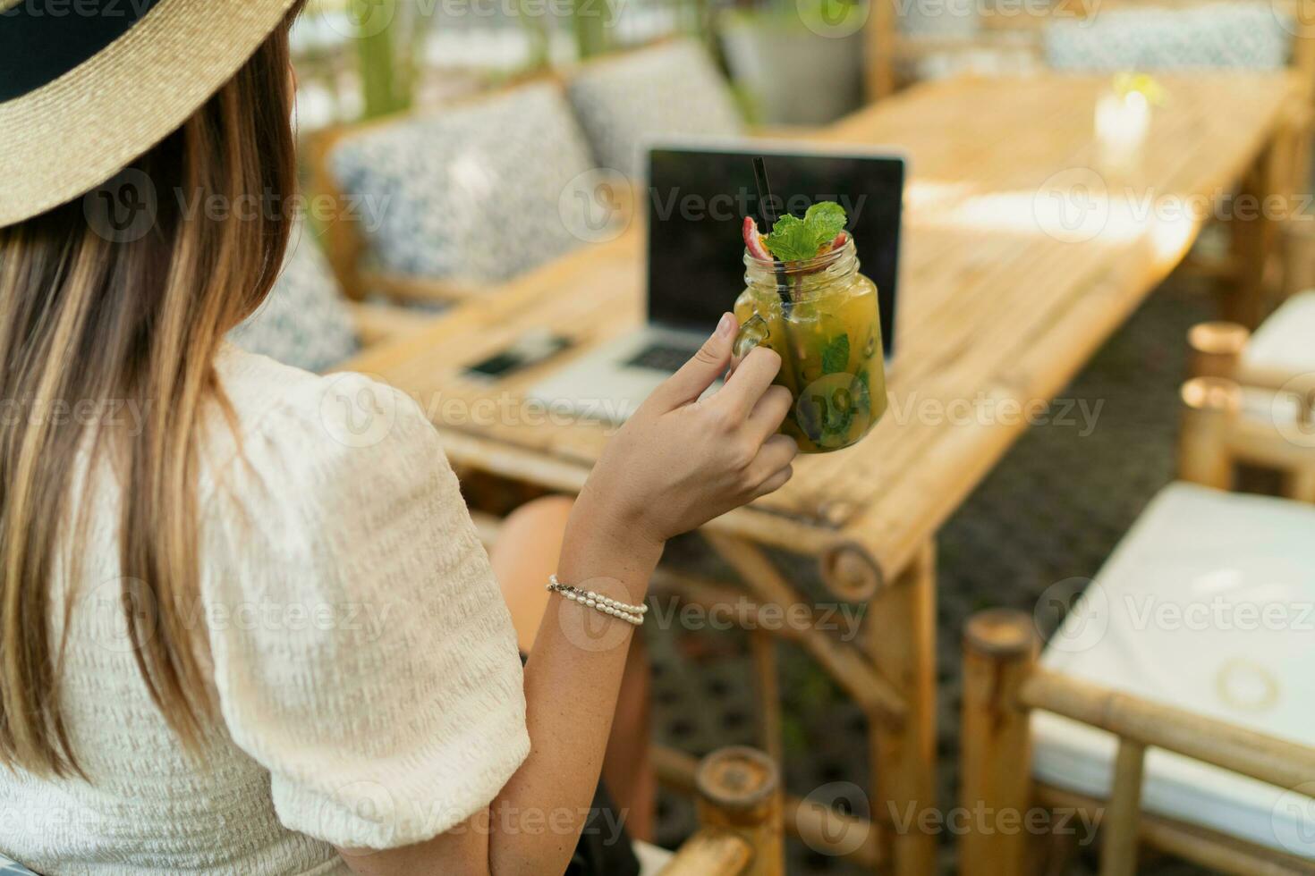 Pretty woman in straw hat using lap top while traveling in asia.  Working remotely in stylish tropical cafe. Holding exotic beverage. photo