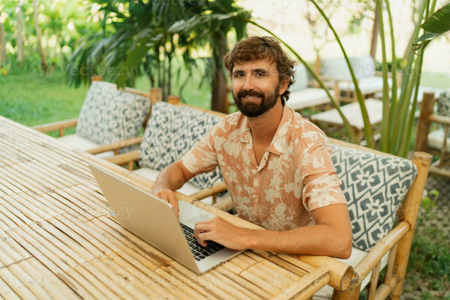 Handsome man with beard using lap top and mobyle phone , sitting in outdoor  cafe with tropical interior. photo