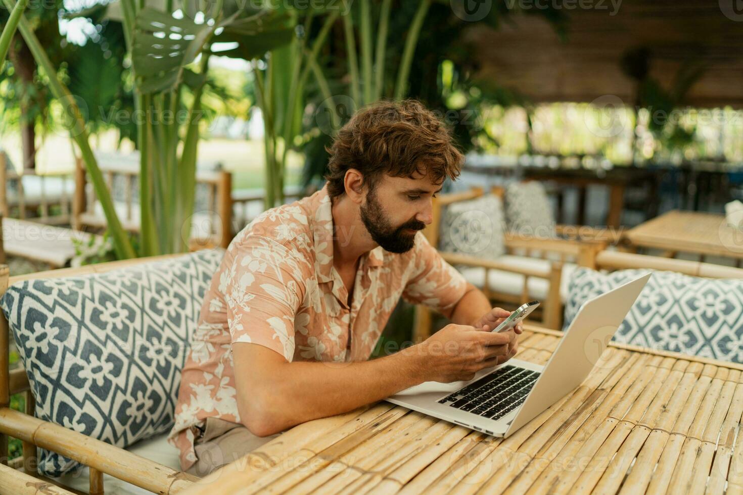 Handsome man with beard using lap top and mobyle phone , sitting in outdoor  cafe with tropical interior. photo