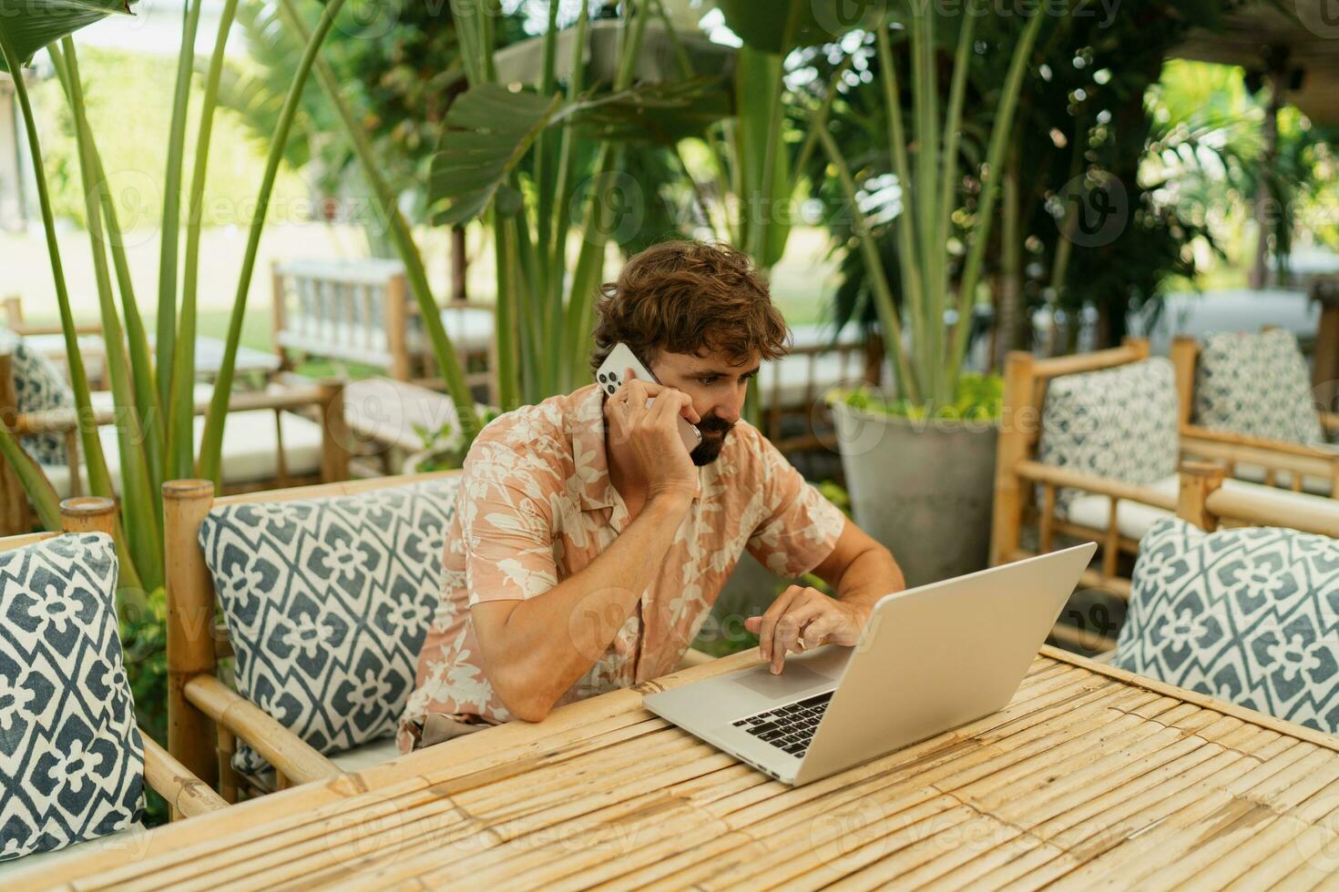 Handsome man with beard using lap top and mobyle phone , sitting in outdoor  cafe with tropical interior. photo