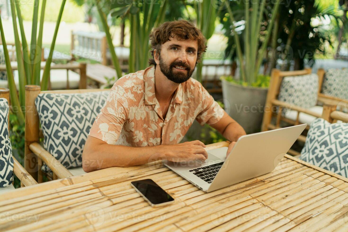 Handsome man with beard using lap top and mobyle phone , sitting in outdoor  cafe with tropical interior. photo
