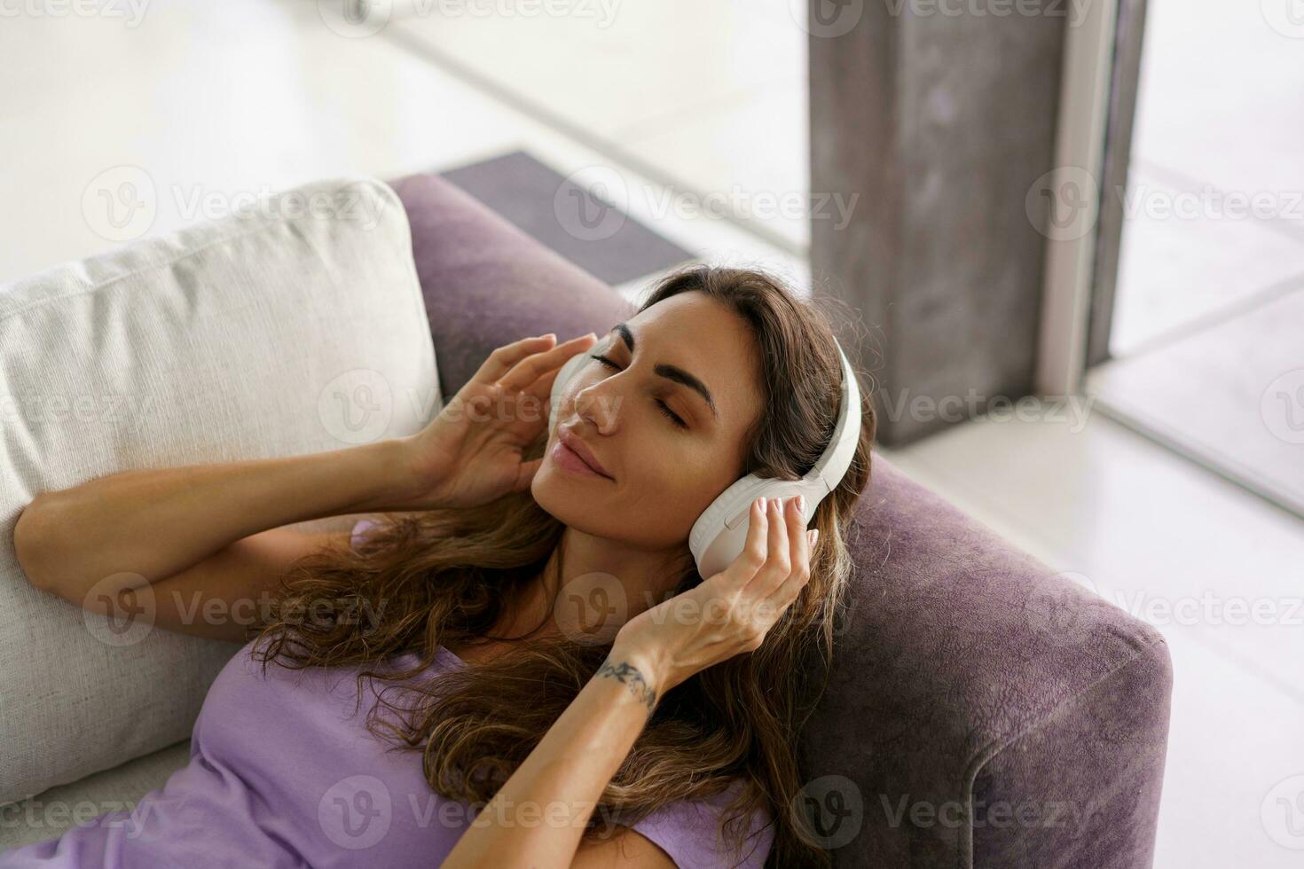 Smiling woman listening to lively music by big white earphones. Enjoying time at home , lieing on sofa. photo
