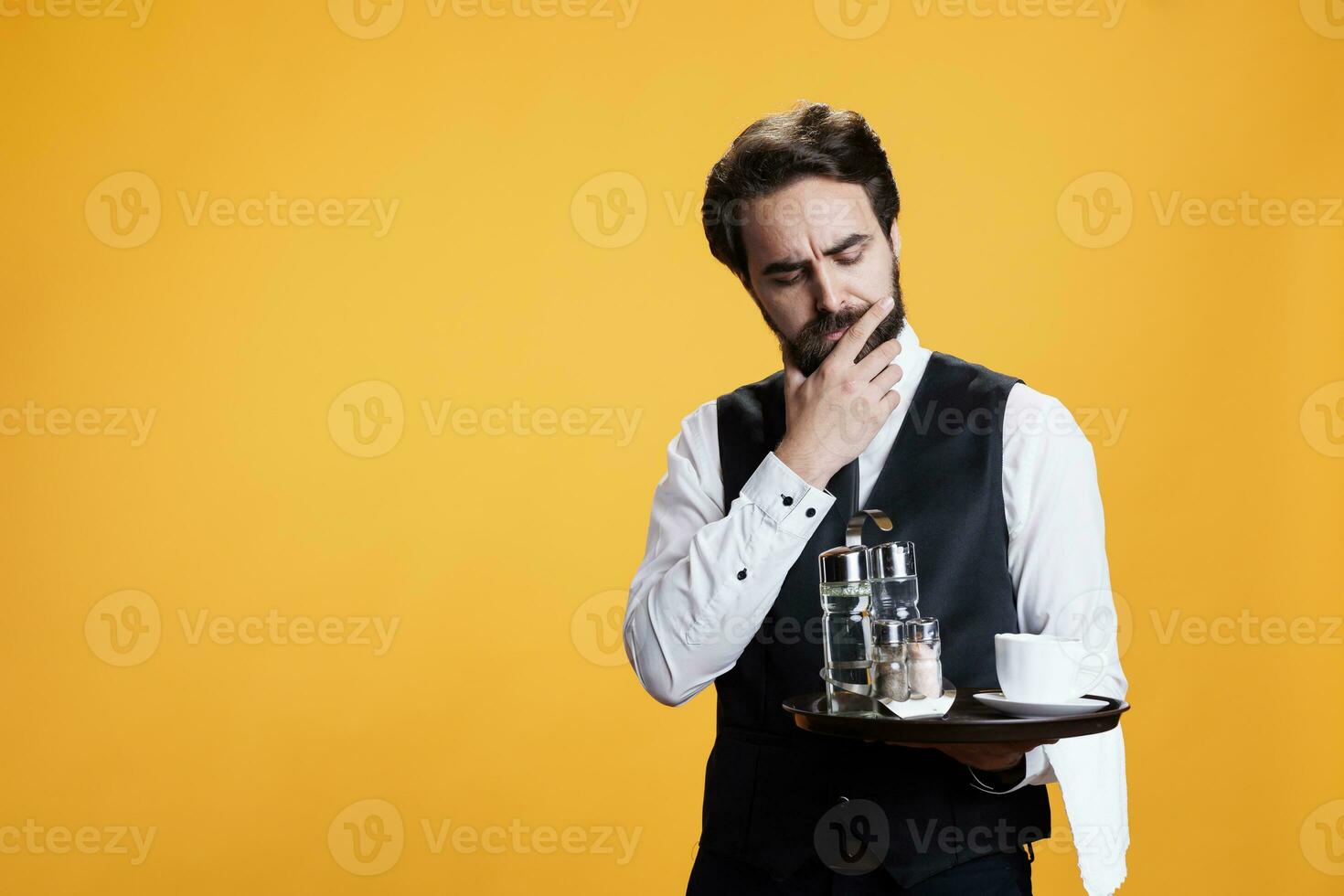 Luxury butler thinking about ideas, preparing to serve customers with food or drinks in yellow studio. Stylish person with waiter occupation being thoughtful with tray in hand on camera. photo