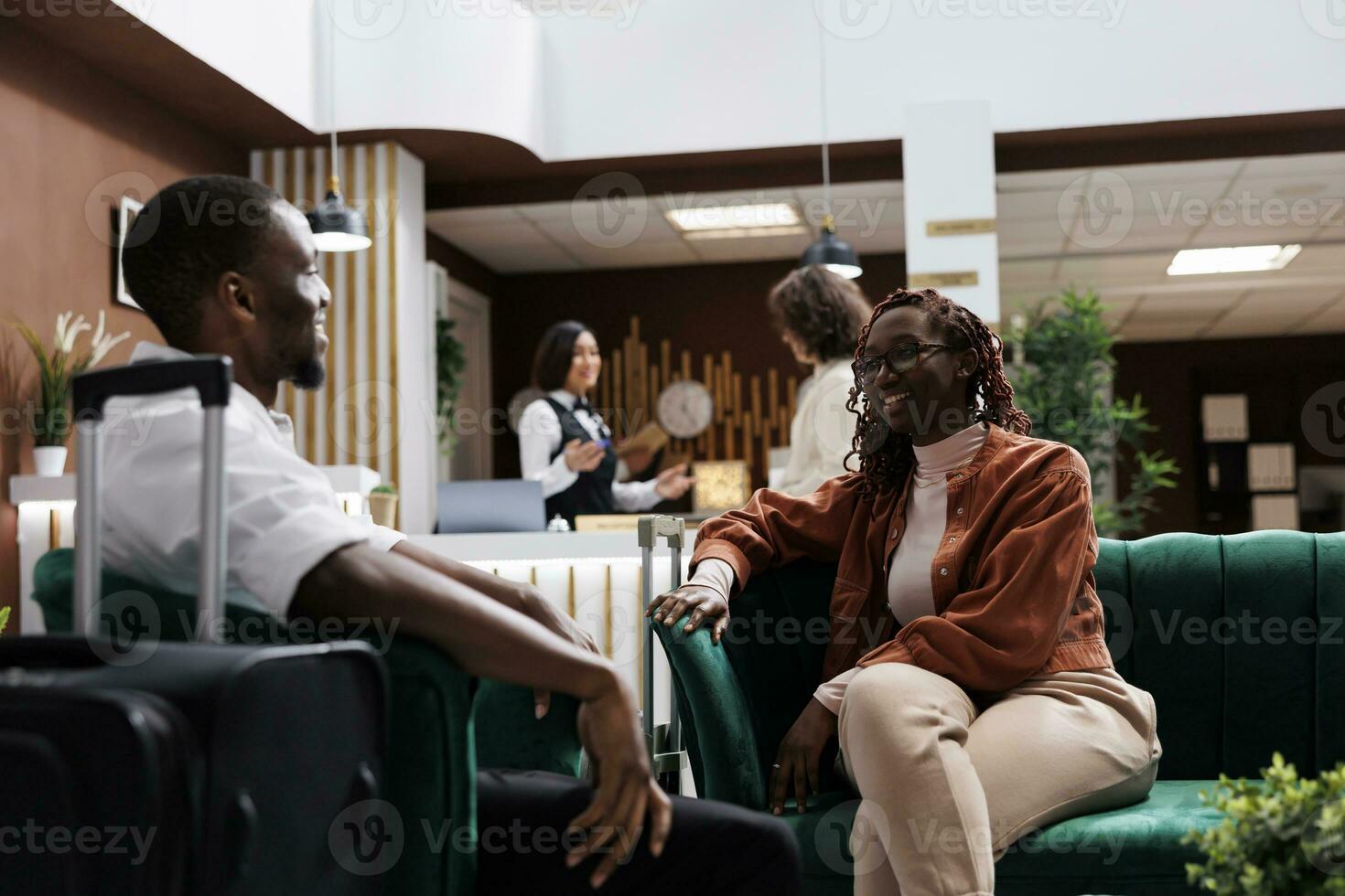 Young couple waiting to do check in at hotel reception, relaxing on couch in luxury lounge area. African american people sitting in modern resort lobby before filling in registration forms. photo