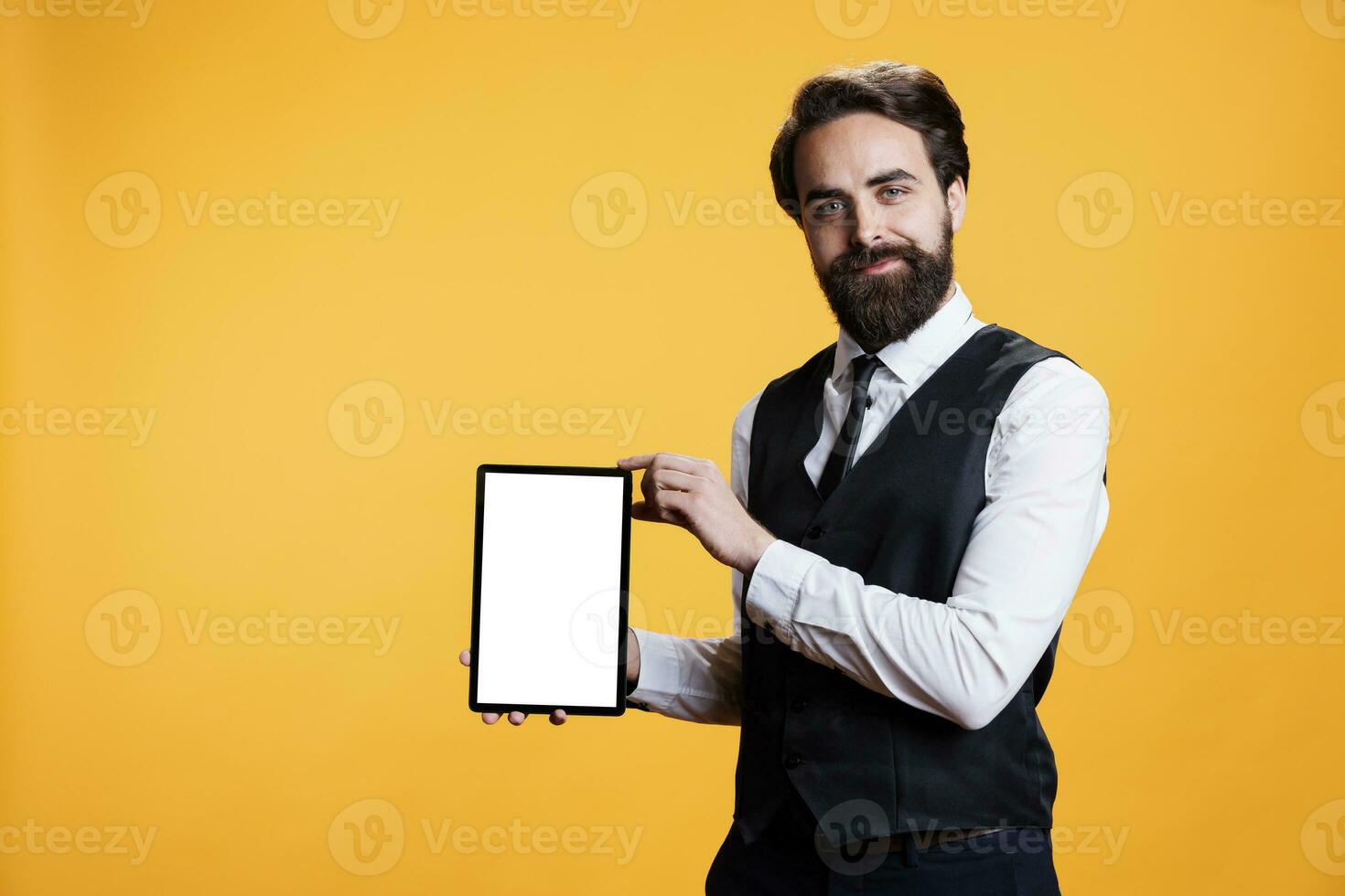 Young waiter holding tablet with white screen on camera, presenting isolated empty template against yellow background. Professional employee shows device with blank mockup display. photo