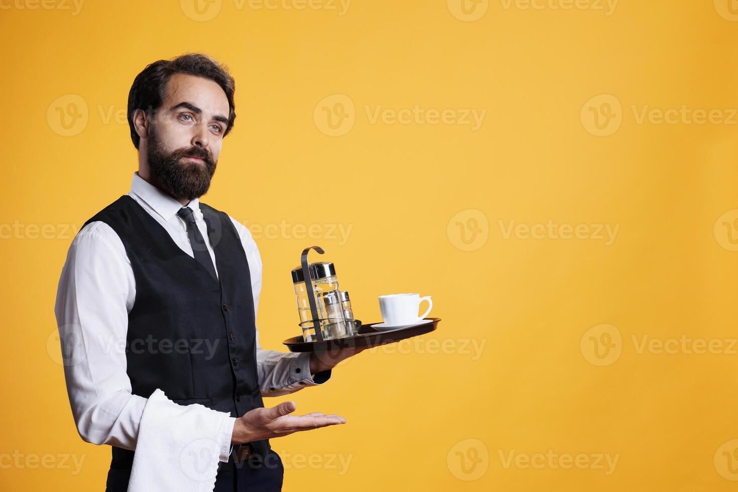 Skilled elegant butler pointing aside and holding food platter to serve customers, working at five star restaurant. Young adult dressed in suit and tie carries tray with cup of coffee. photo
