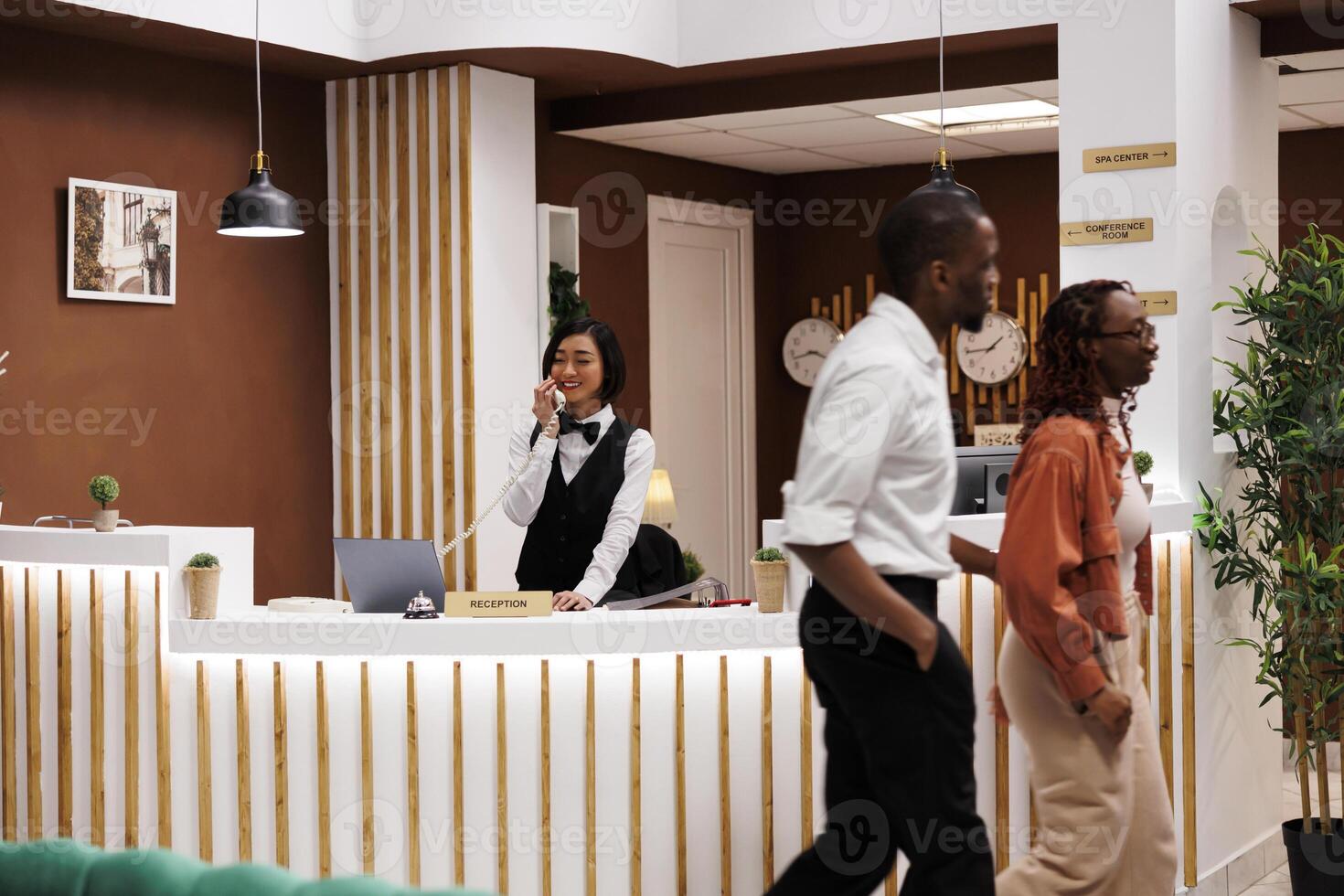 Asian receptionist making online booking using landline phone at reception counter front desk, talking to people about room reservations. Hotel staff working on welcoming tourists. photo