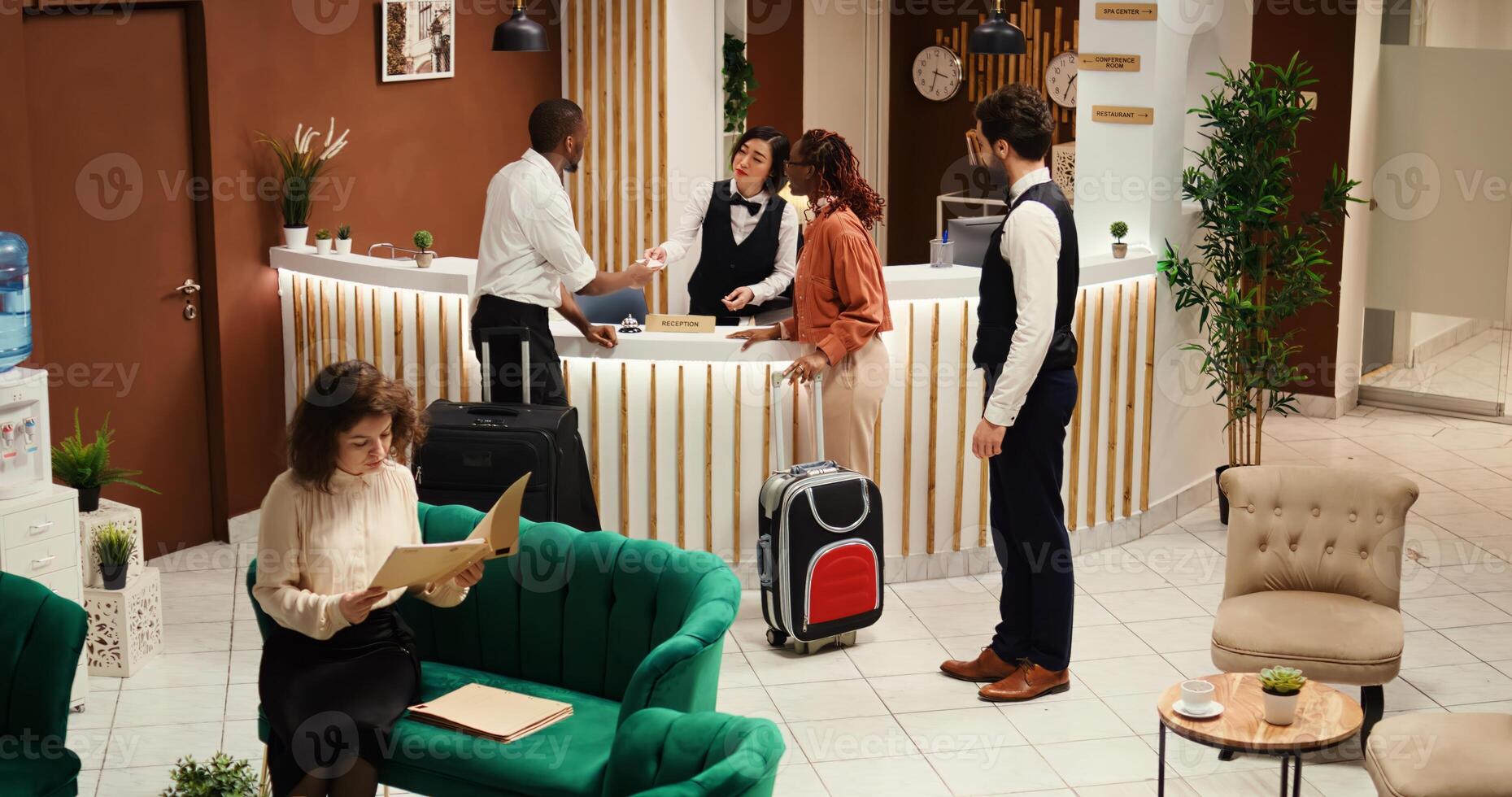 Focused hotel manager verifying tourists booking paperwork while they are being assisted by concierge personnel. Bellboy helping guests with baggage after check in process is over. photo