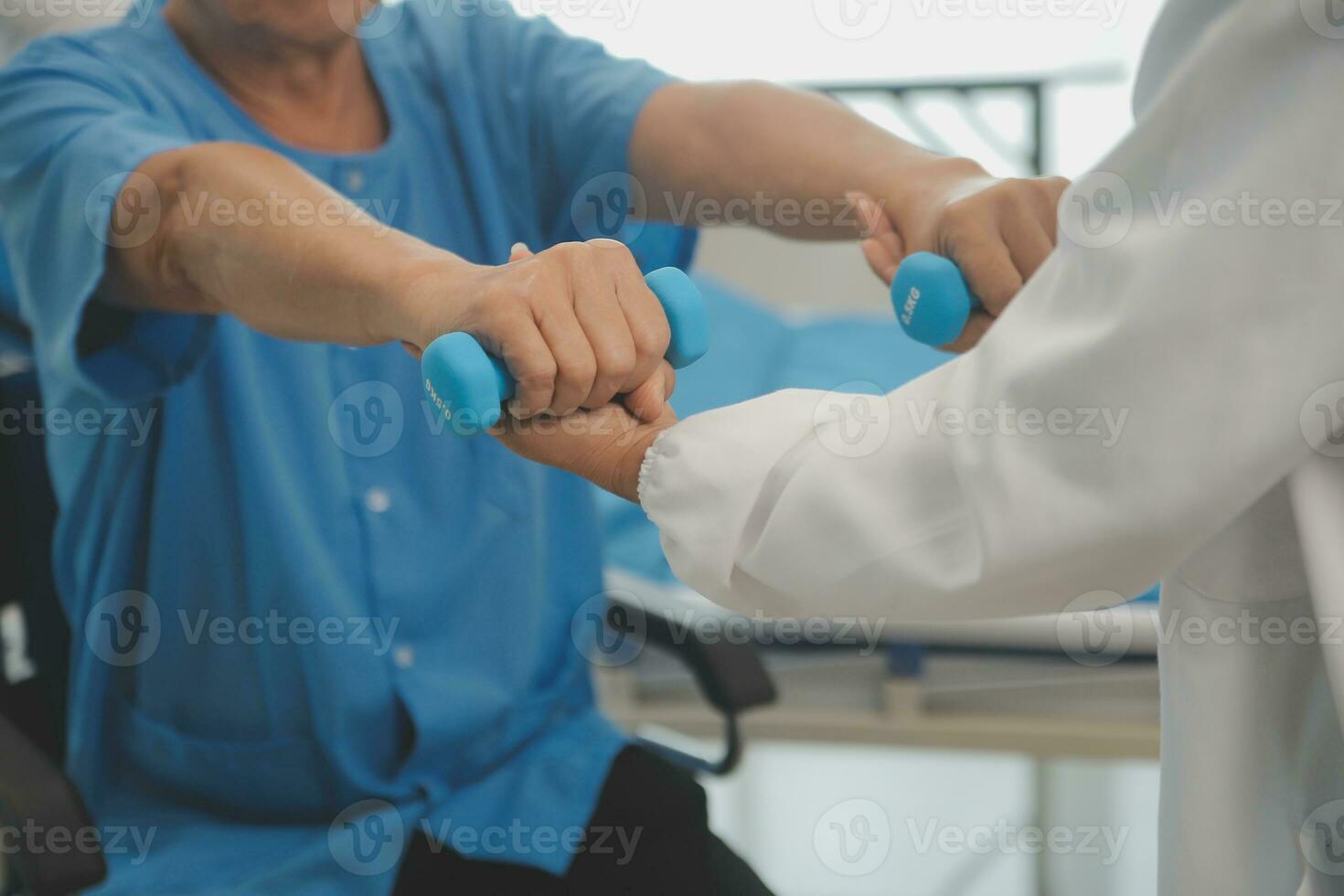 Asian older aged man doing physiotherapist with support from nurse. Senior elderly male sitting on sofa in living room using dumbbells workout exercise for patient with caregiver in nursing care. photo