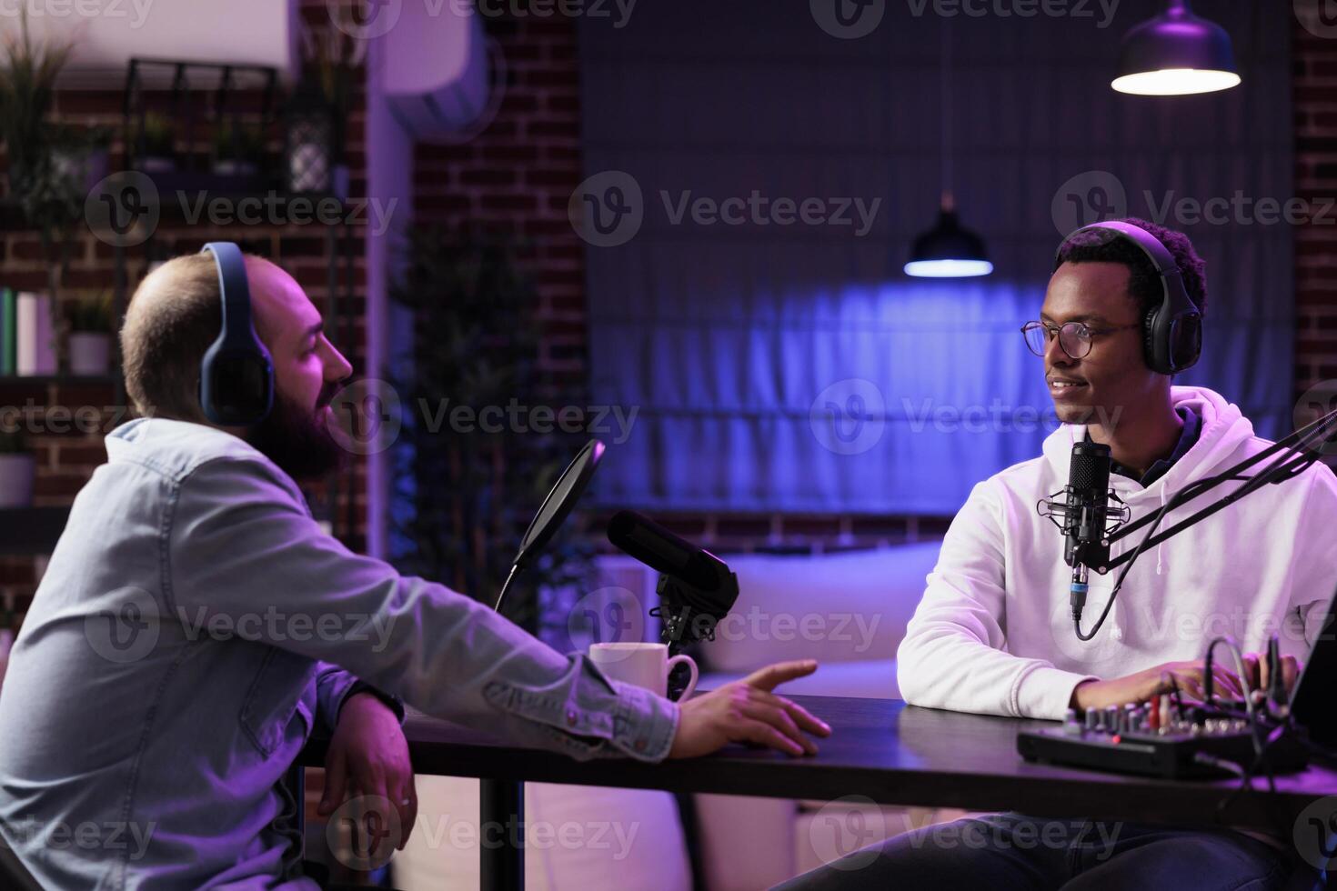 Influencer recording podcast with guest, using audio recording equipment in home studio living room. Cheerful men chatting live during broadcasting session for online streaming service show photo