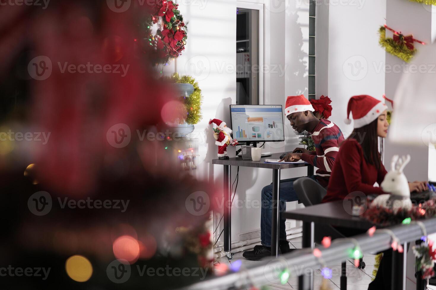Focused african american businessman solving tasks on computer sitting at desk during holiday season. Meticulous employee wearing christmas hat, working in xmas decorated workplace office photo