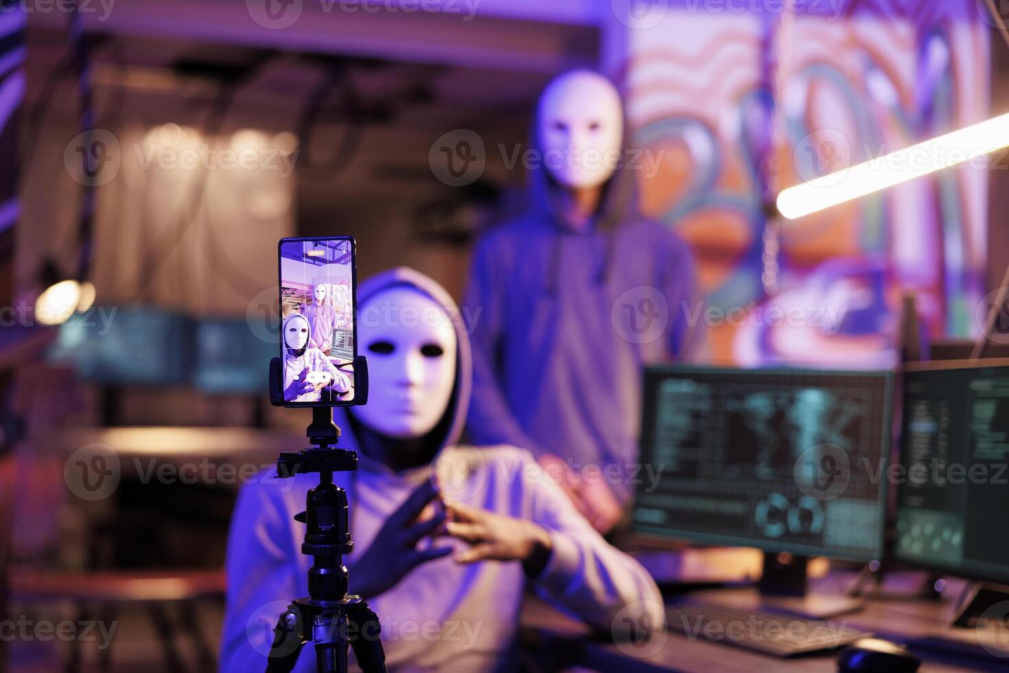 Mobile phone screen displaying anonymous hacker recording scam message. Internet criminals in masks and hoods live streaming on smartphone in abandoned warehouse at night photo