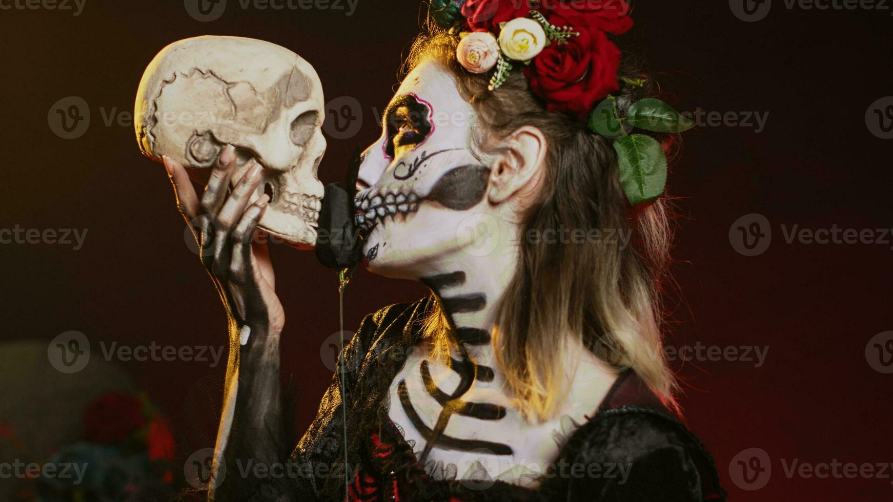 Flirty dead lady acting creepy with skull in hand, holding black roses and wearing scary make up. Looking like santa muerte with body art on holy mexican celebration, being charming. Handheld shot. photo