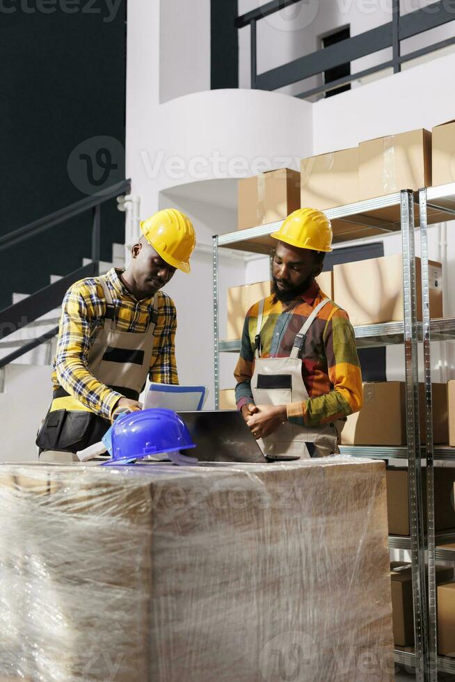 Logistics managers analyzing inventory and comparing data on laptop and clipboard in warehouse. African american men checking goods supply schedule and working in distribution department storehouse photo