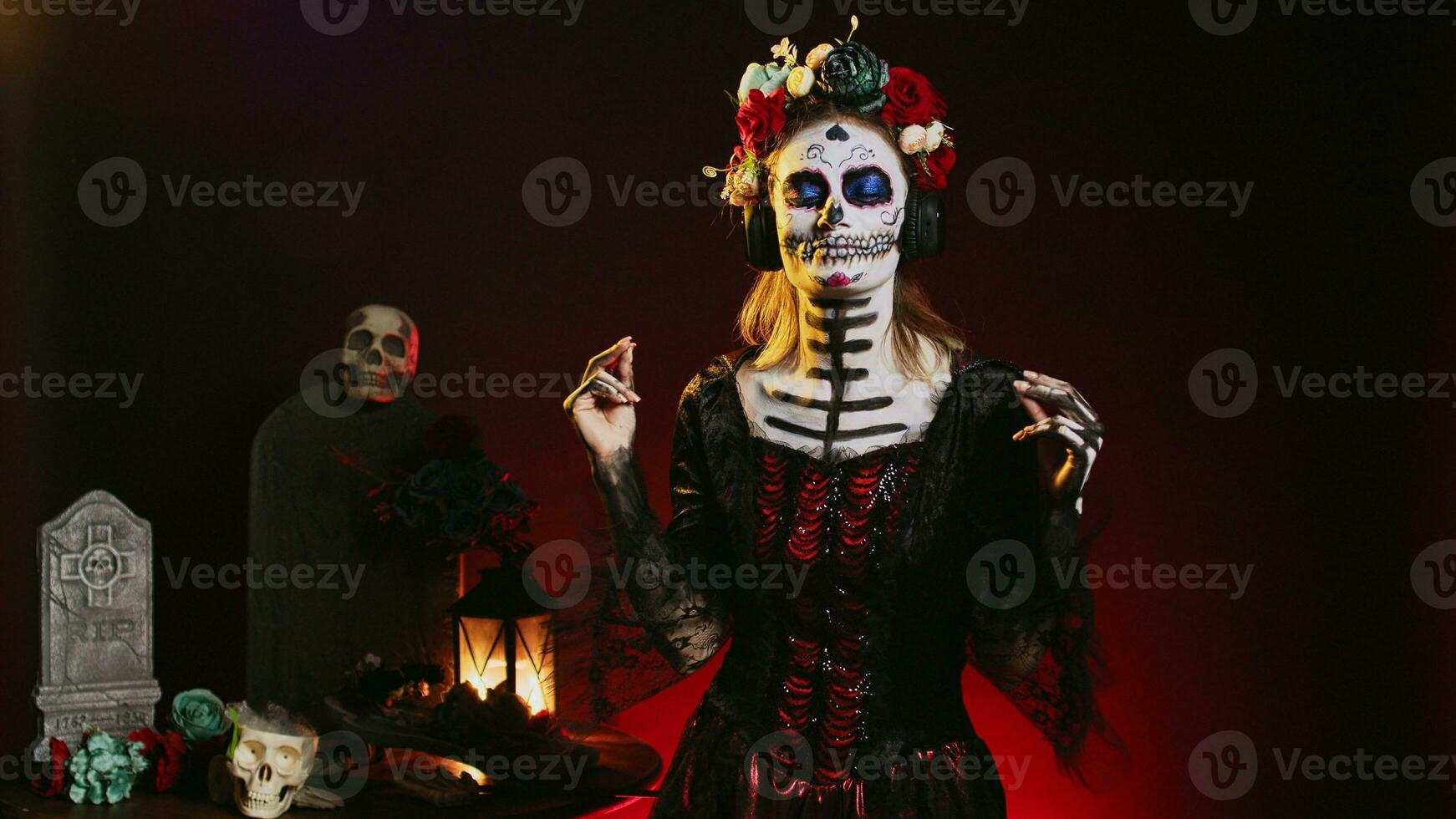 Goddess of dead enjoying music on headphones, wearing skull make up and santa muerte costume in studio. Beautiful person listening to song on audio headset, celebrating day of the dead. photo