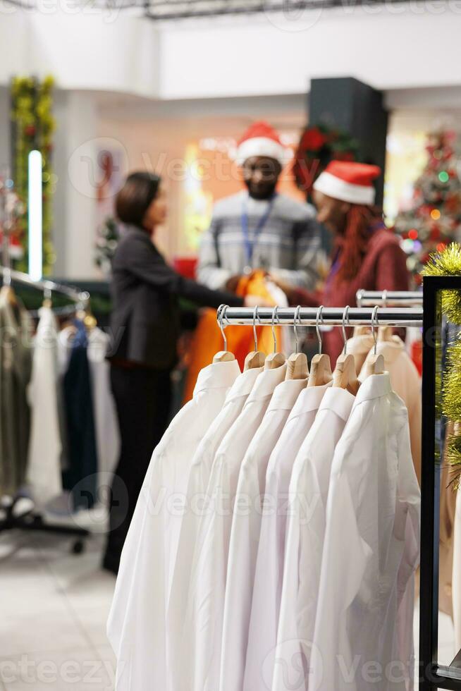 Selective focus of staff helps client in clothing store, presenting new clothing line models to buy on sale. Retail employees assisting customer to choose festive dinner outfit for christmas eve. photo