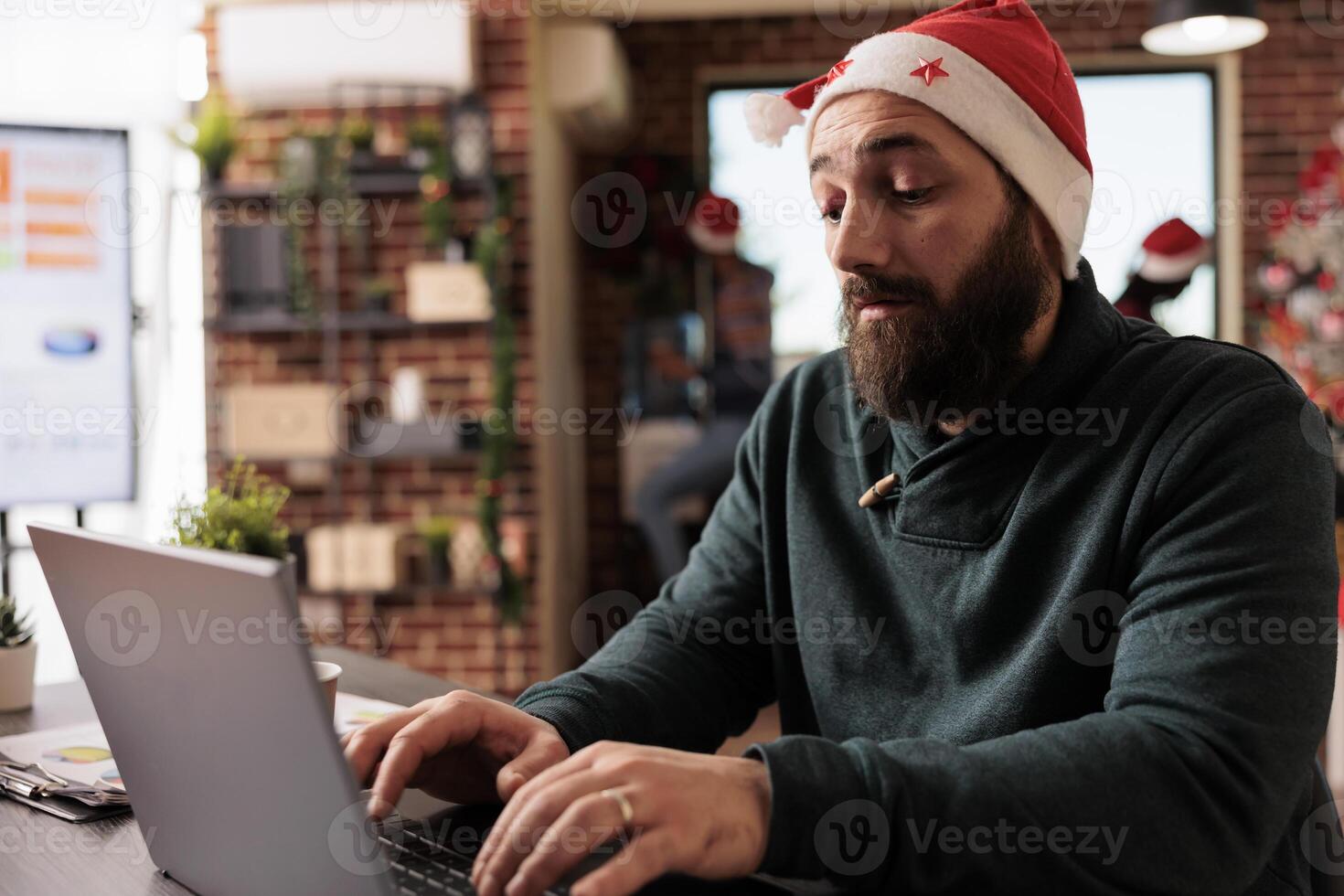 Startup company worker typing message on laptop while working in office at christmas eve. Corporate employee wearing santa claus hat using computer and sitting at workplace desk photo