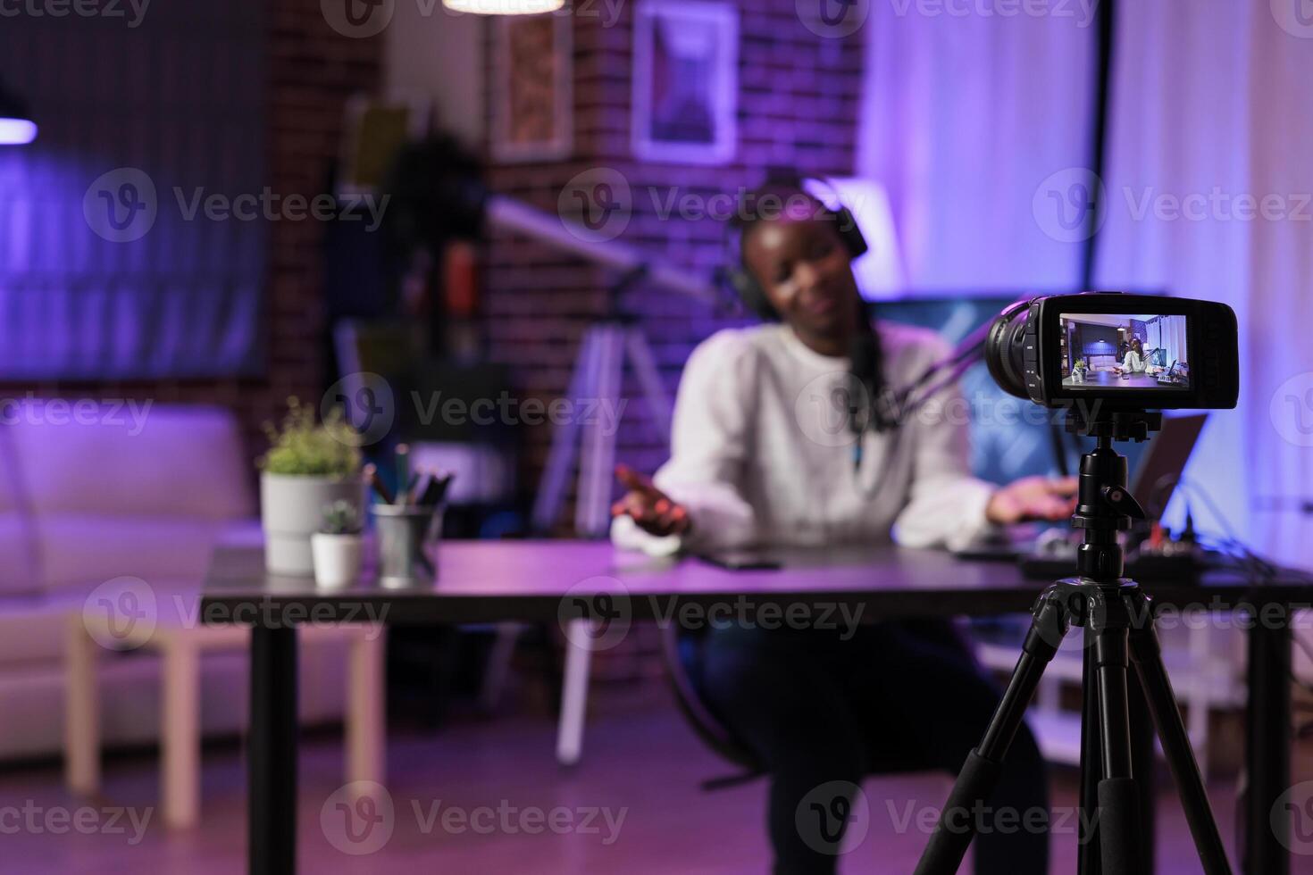 Focus shot on modern camera recording vlogger in blurry background talking about parenting subjects. Woman filming online show using professional gadgets in rgb lights studio photo