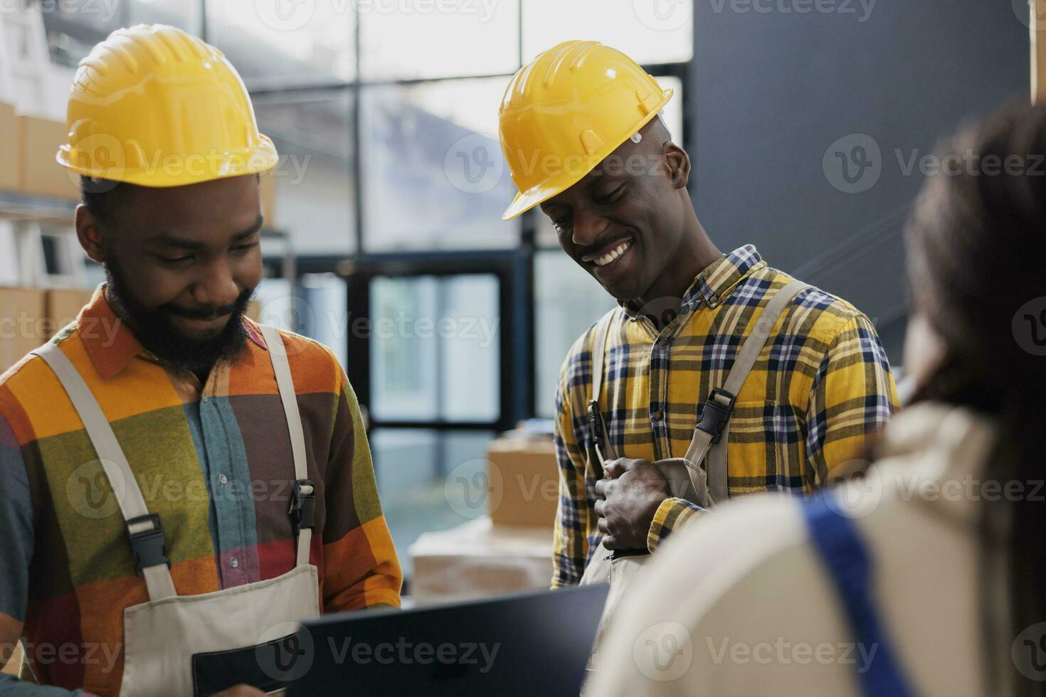 Warehouse workers wearing yellow helmets laughing and chatting at work. Smiling african american men storehouse employees in protective hard hats and overall having fun conversation photo