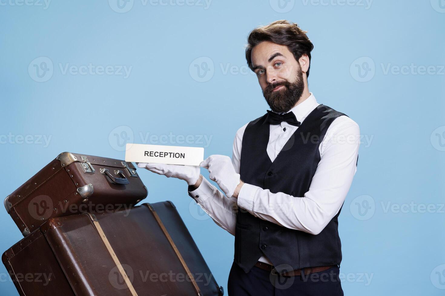 Bellhop shows reception indicator on camera, pointing guests in the right direction. Professional hotel doorkeeper working in tourism industry, holding front desk pointer in studio. photo