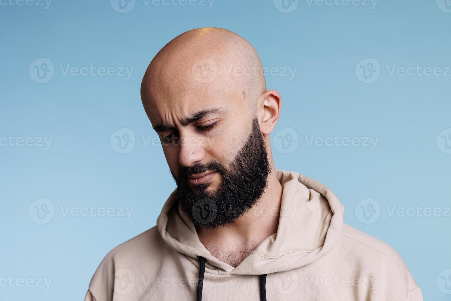 Thoughtful arab man tilting head down and looking away. Young bald bearded person thinking with uncertain facial expression and posing casually in studio on blue background photo