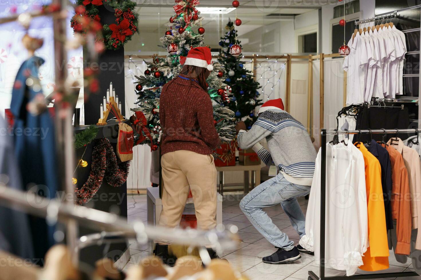 Al por menor trabajadores decorando Navidad árbol en ropa Tienda a compras centro, festivo empleados vistiendo Papa Noel sombreros y colgando guirnaldas personal miembros equipo preparando para fiesta estación. foto