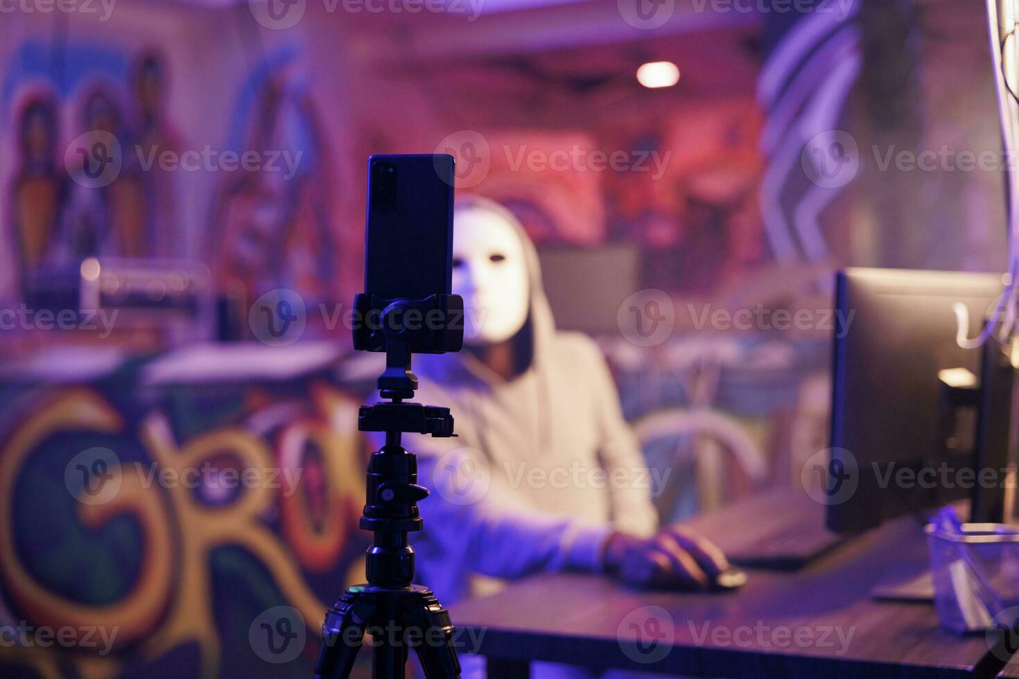 Anonymous criminal recording scam on smartphone camera in abandoned warehouse. Hacker with hidden identity doing illegal activity while filming threat on mobile phone on tripod photo